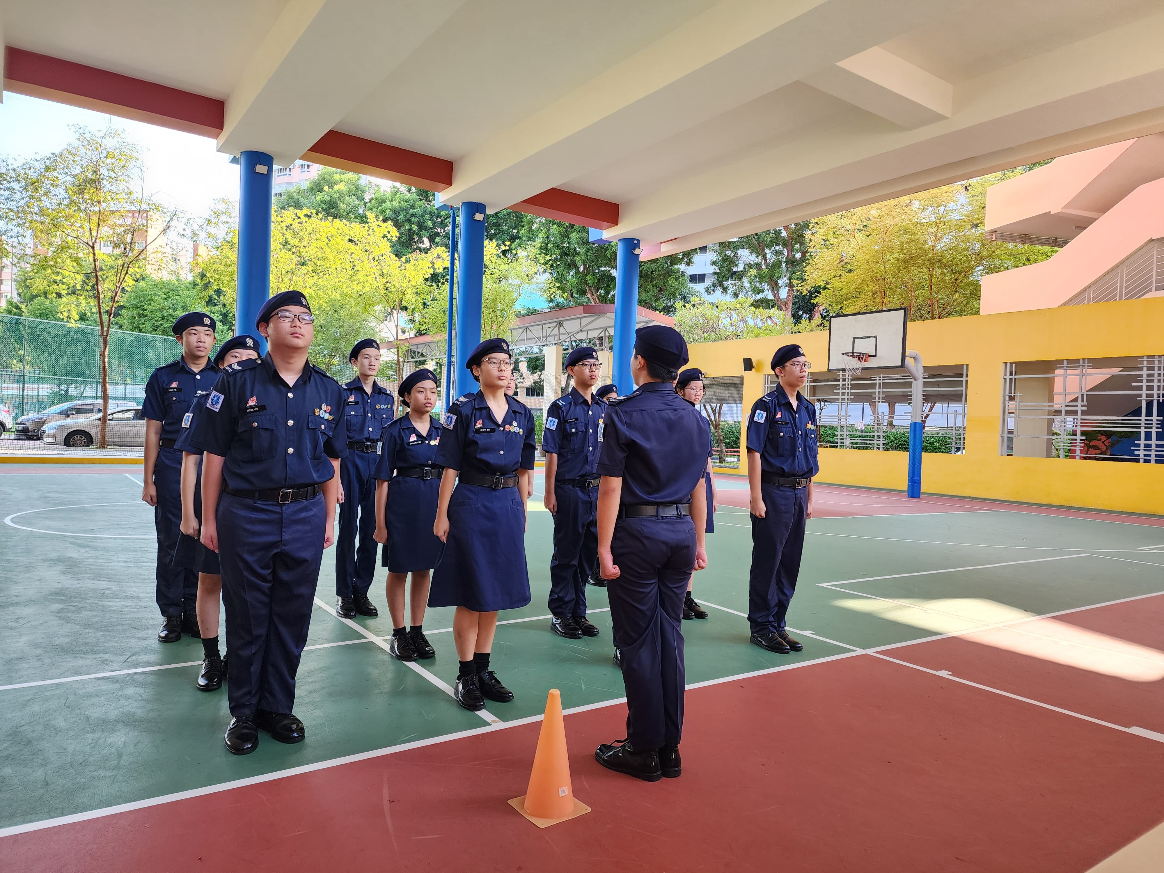 Youth cadets in navy blue uniforms and berets stand at attention on a school court during a drill inspection.