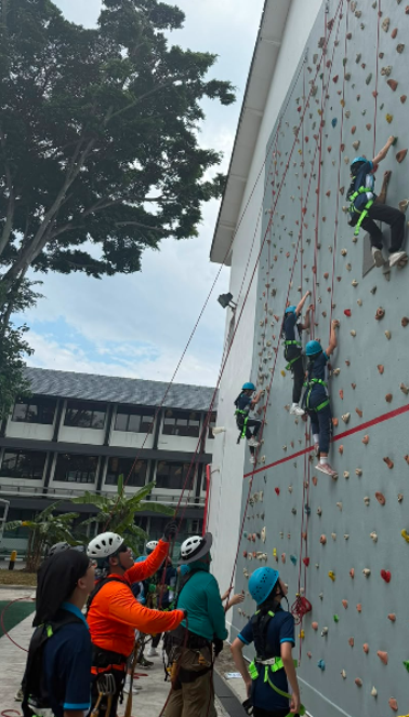 Students in safety helmets and harnesses outdoor rock climbing on a school climbing wall while classmates belay from below.