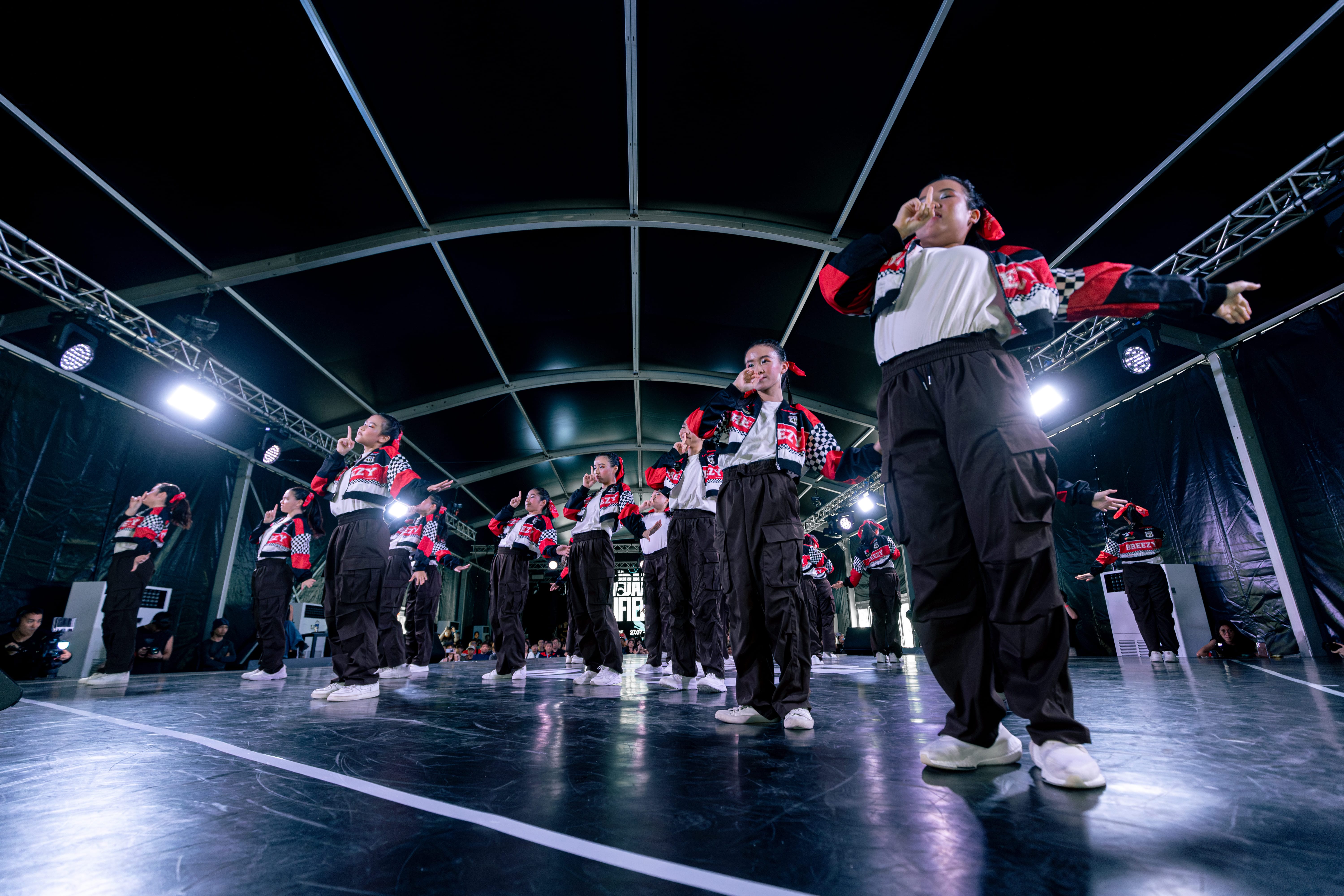 A dance crew in matching red, black, and white jackets with black cargo pants performs on a lit indoor stage.