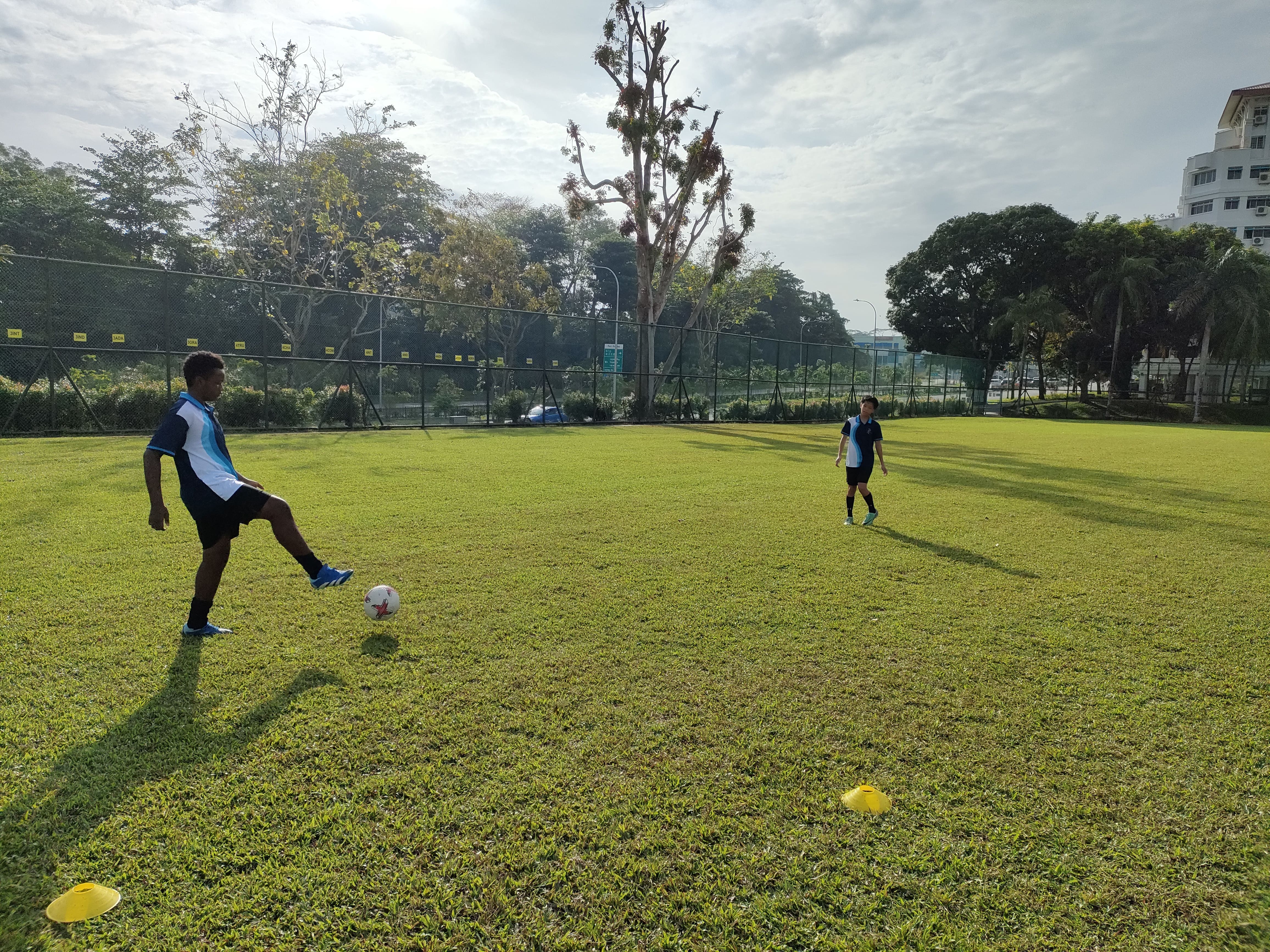 Two players in matching blue jerseys practice passing a football on a green grass field with yellow cones.