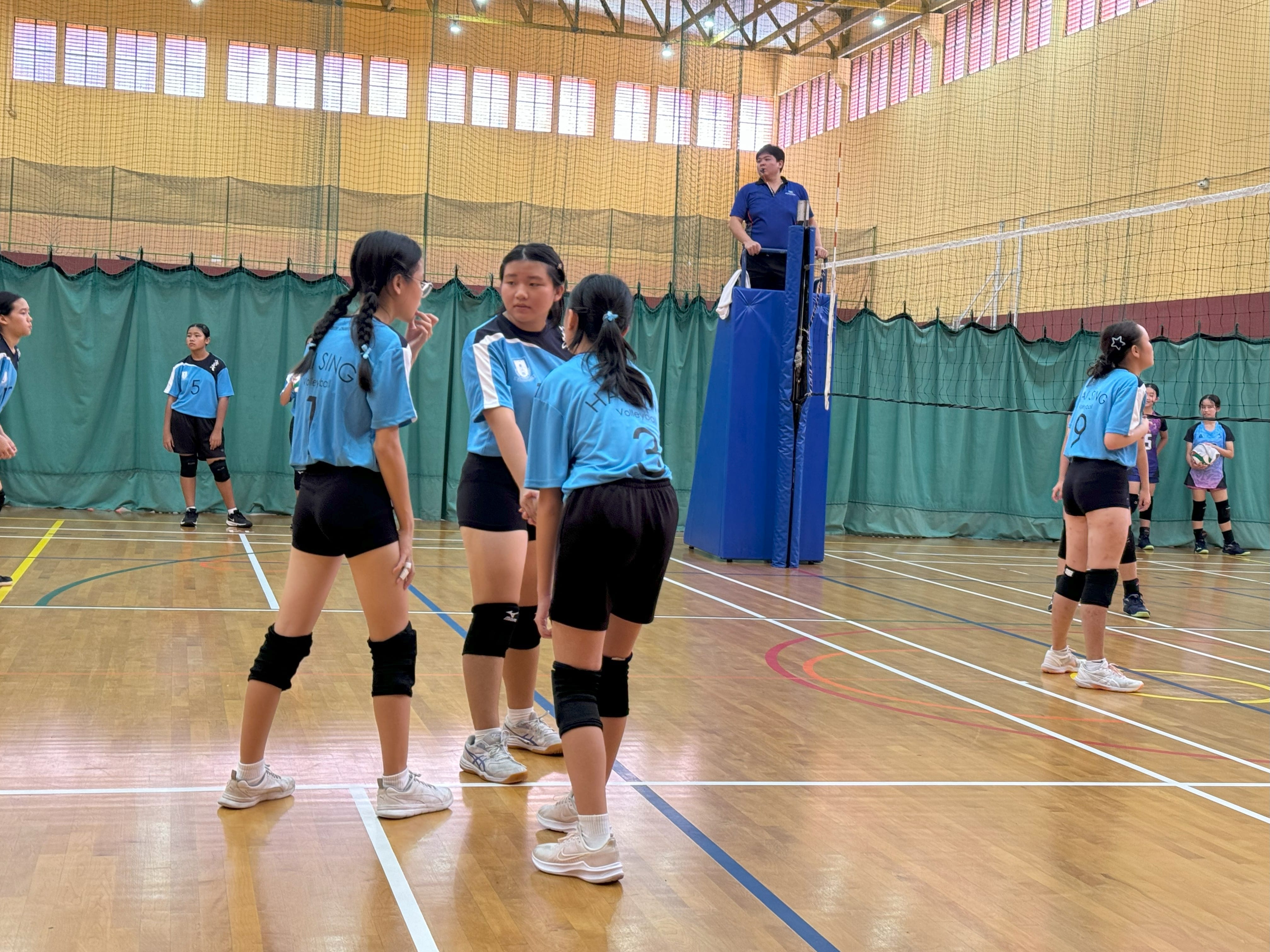 Young girls in blue volleyball uniforms huddle on an indoor court near the net while a referee stands on a platform.