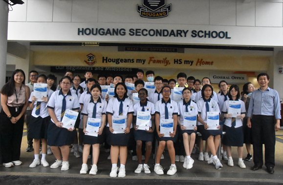 Students in uniforms holding certificates pose in front of Hougang Secondary School entrance.