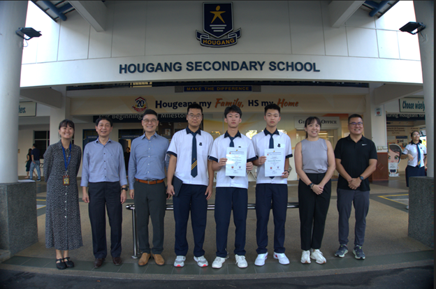 Seven people stand in front of Hougang Secondary School entrance, two students holding certificates.