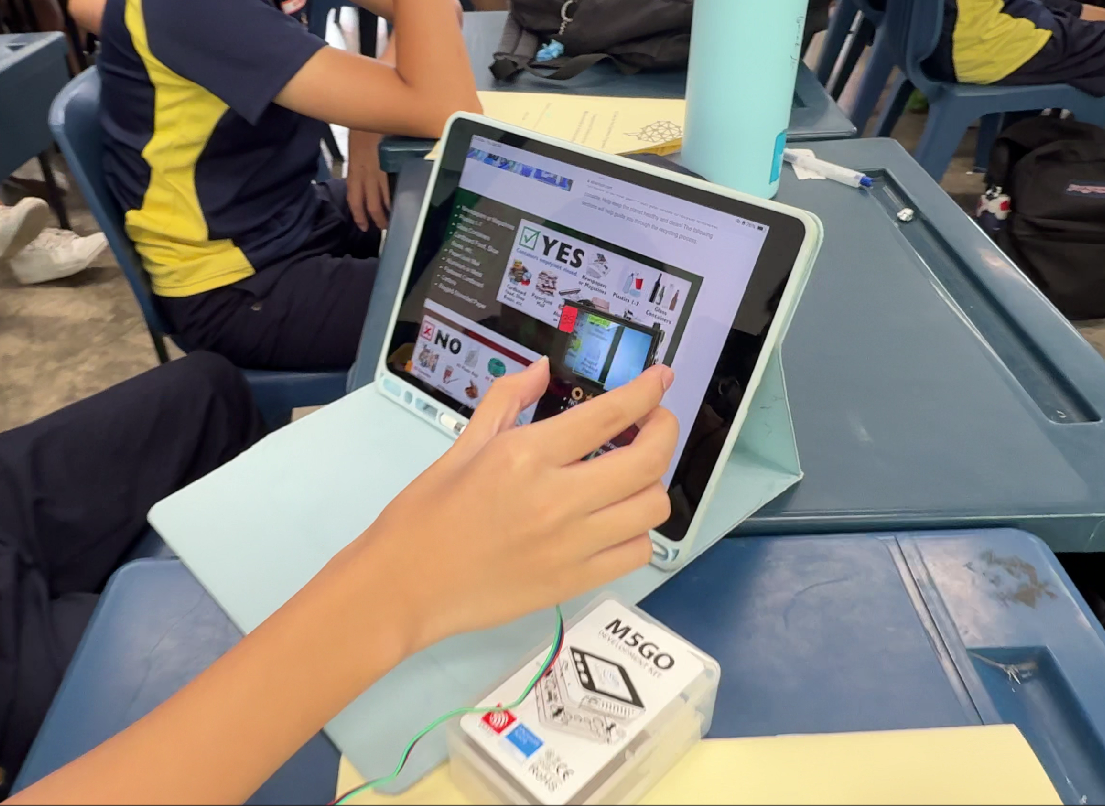 Student's hands holding a tablet showing YES and NO recycling guide, with an M5GO device on a school desk.