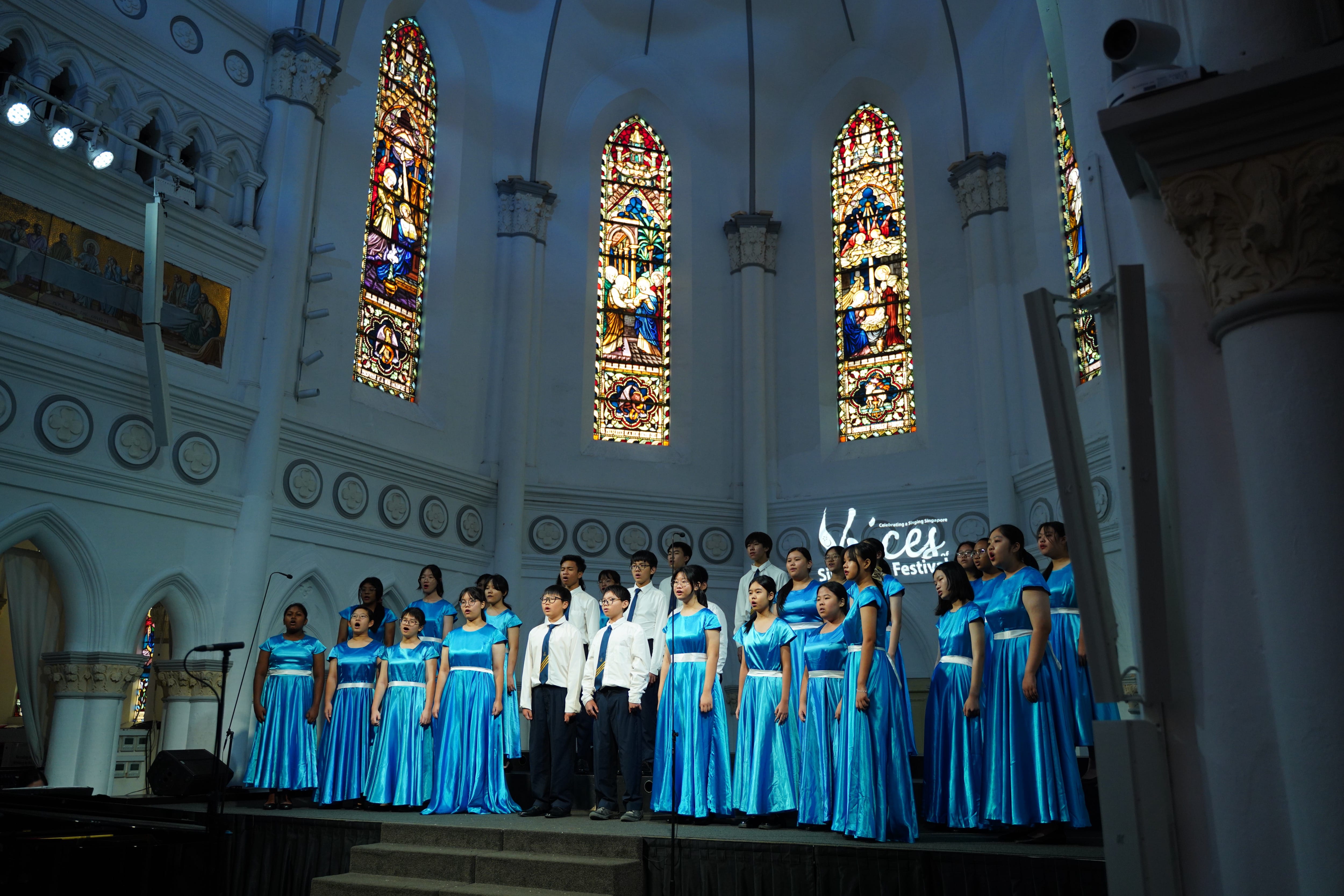 A youth choir in blue satin dresses performs on a church stage with colorful stained glass windows behind them.