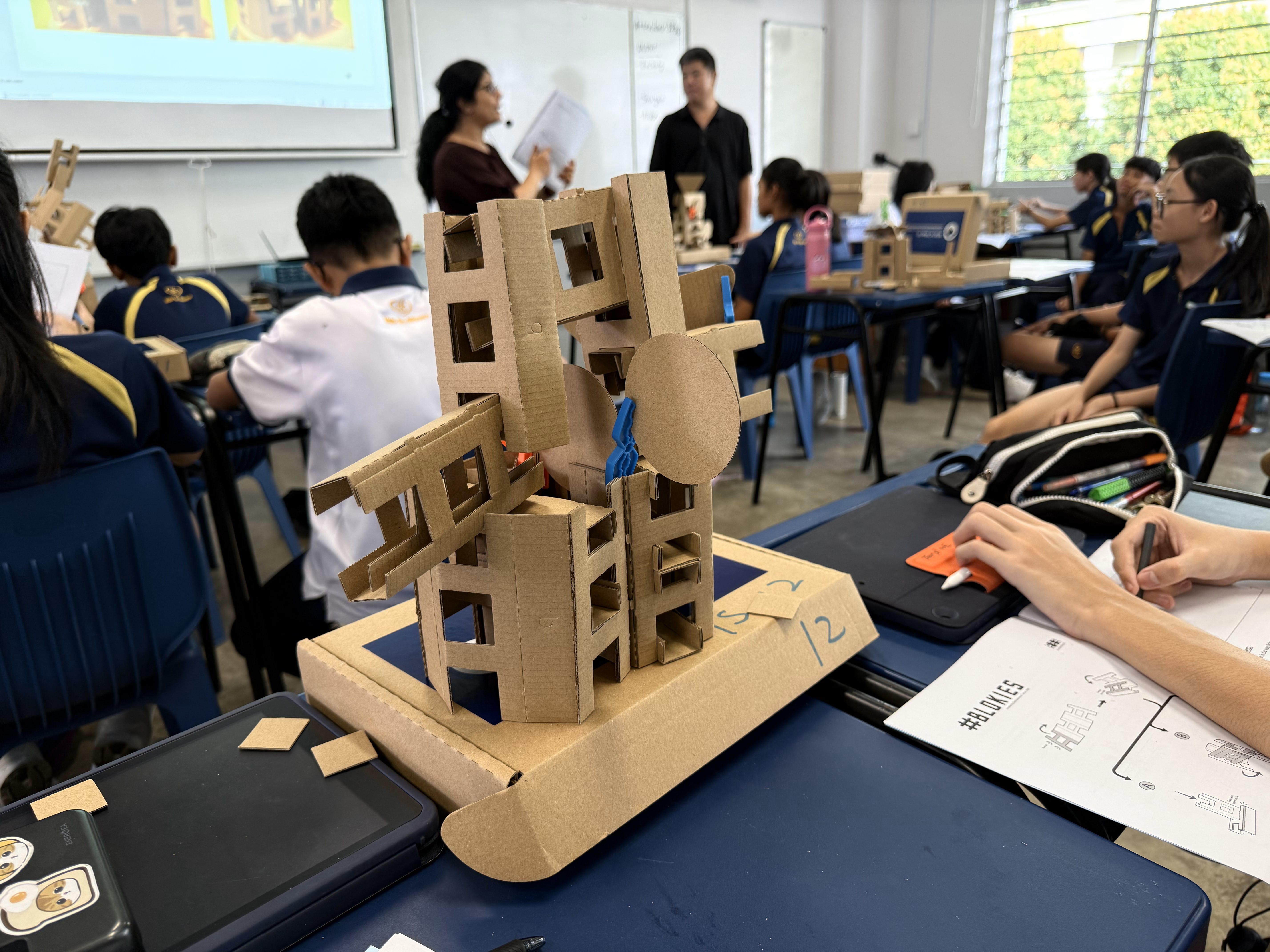 Students in a classroom work on cardboard structural models while two teachers stand near a projector screen.