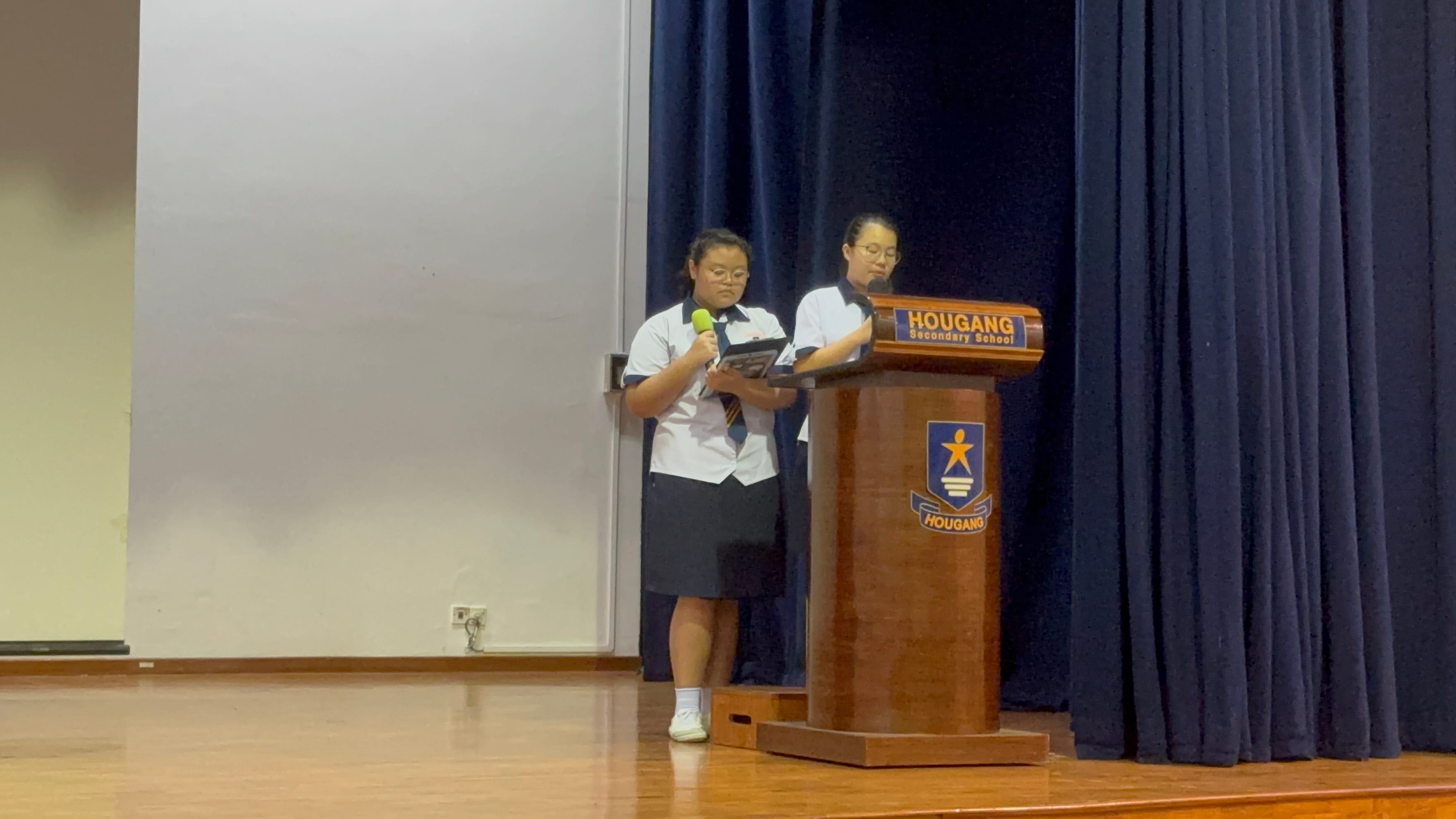 Two uniformed students on stage, one holding a microphone, beside a Hougang Secondary School wooden podium.