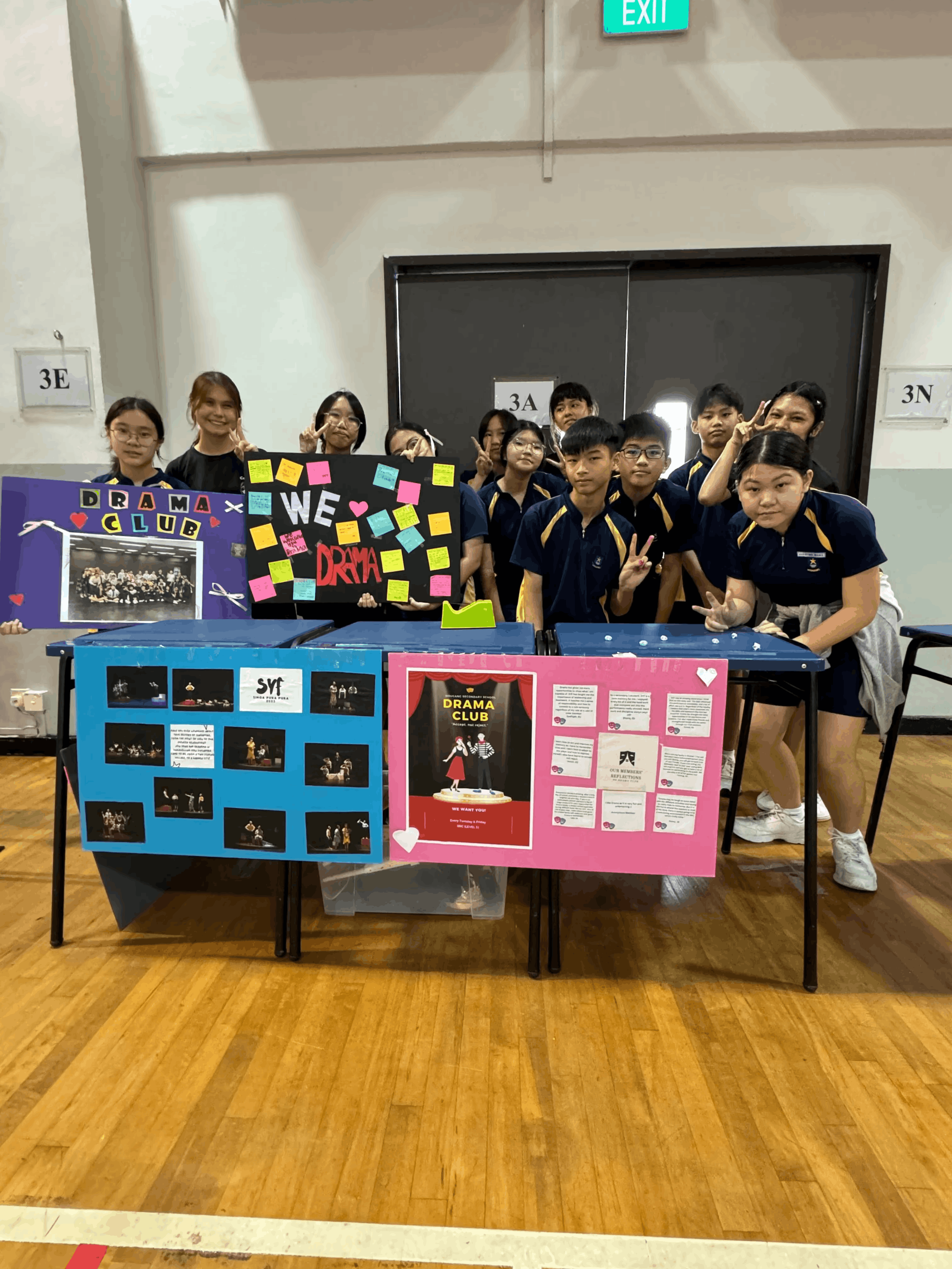 Students in navy uniforms pose behind a Drama Club booth with colorful posters and photo displays in a school hall.