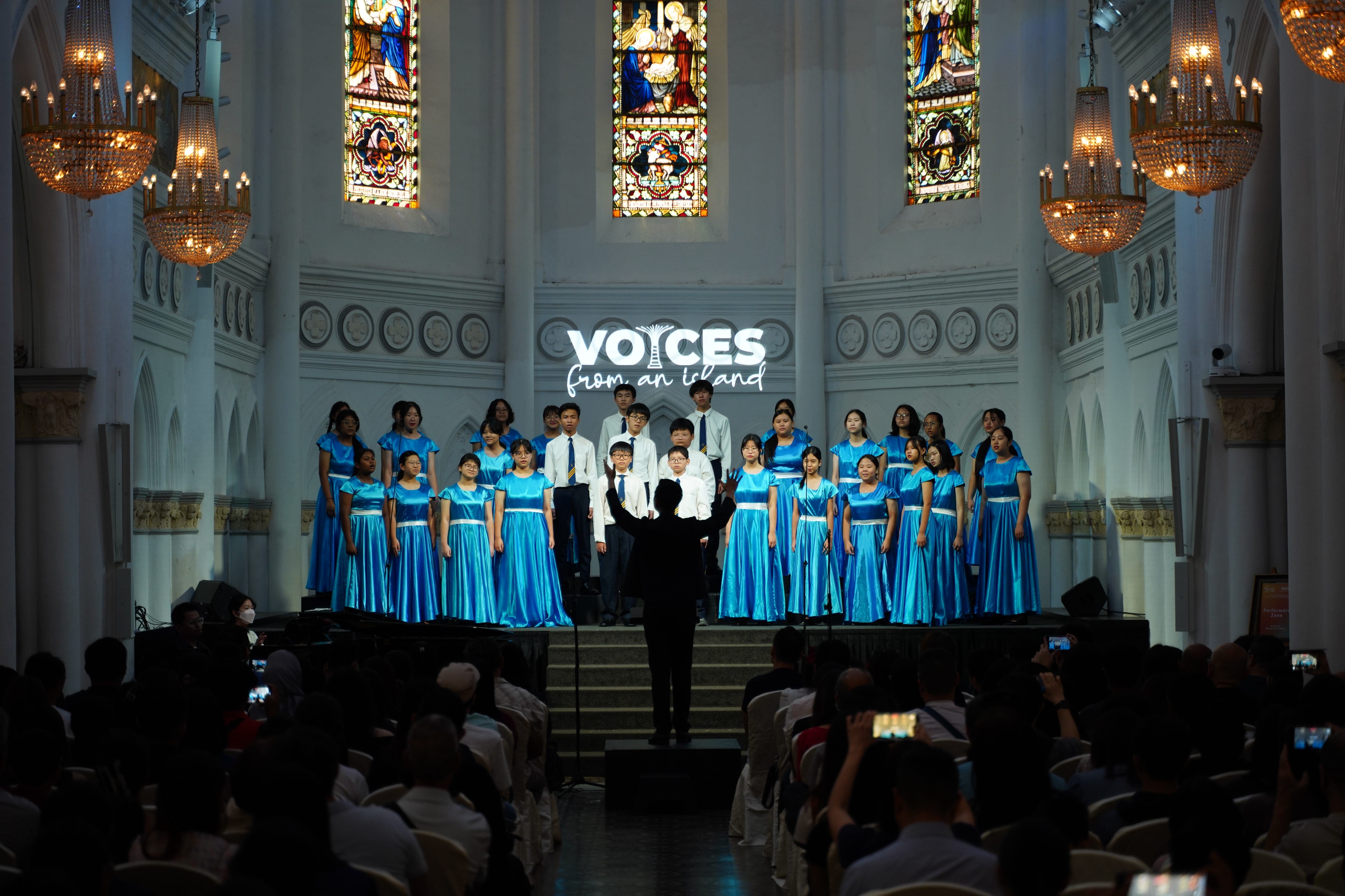 A youth choir in blue satin dresses performs in a church with stained glass windows under the banner "Voices from an Island."