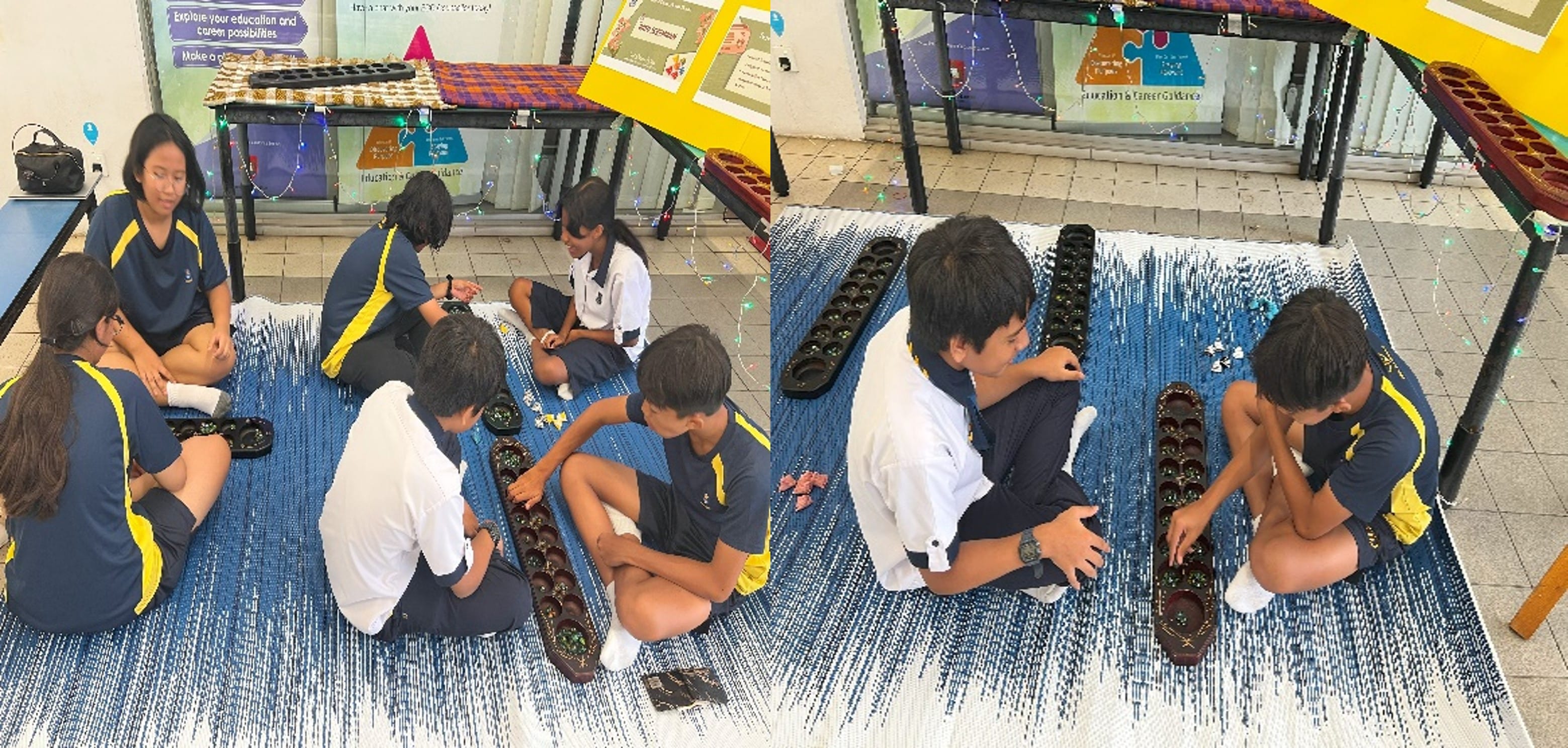 Students in blue and yellow uniforms sit on a mat playing mancala board games outdoors near display boards.