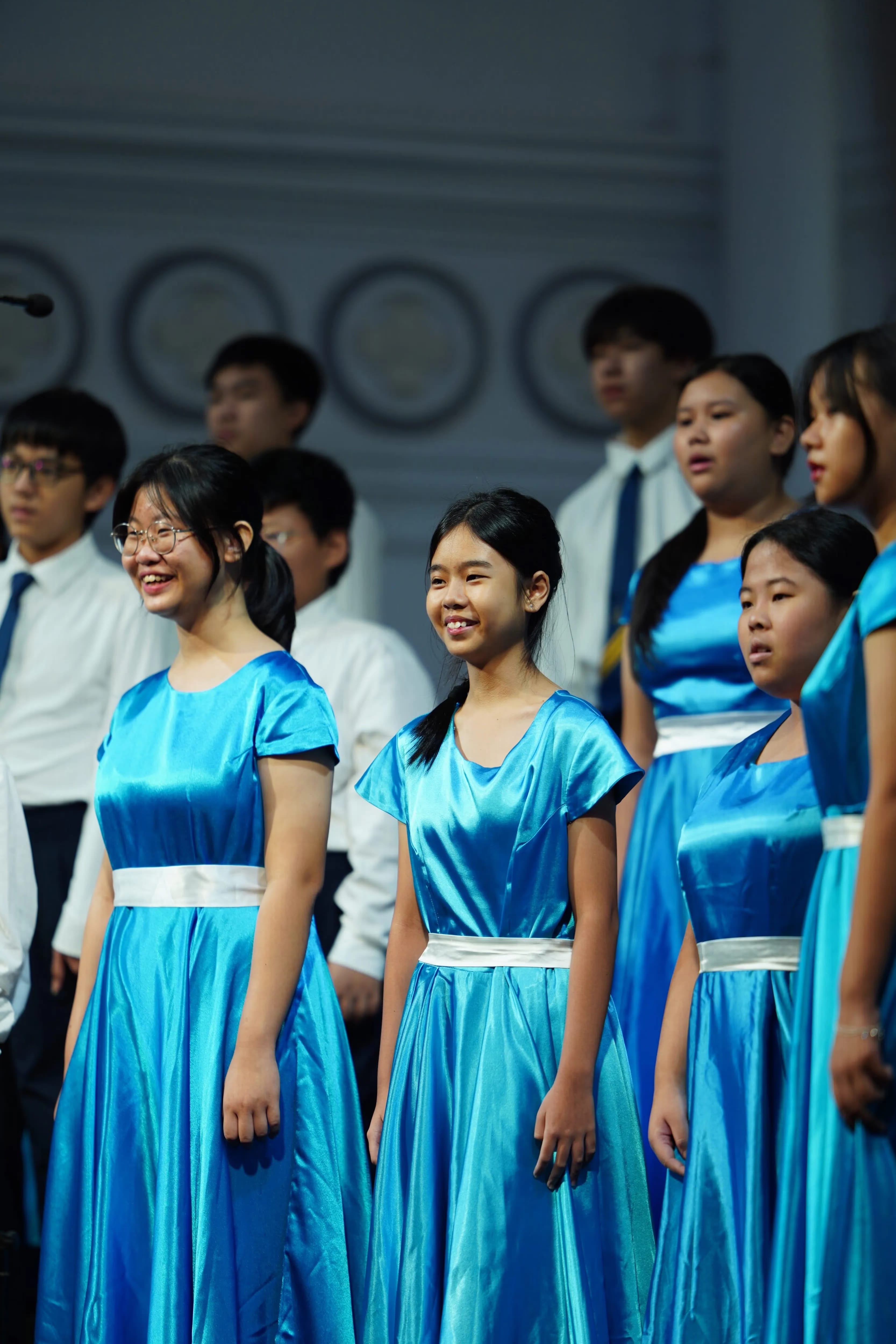 Young choir members perform on stage, girls in blue satin gowns with white sashes, boys in white shirts and ties.
