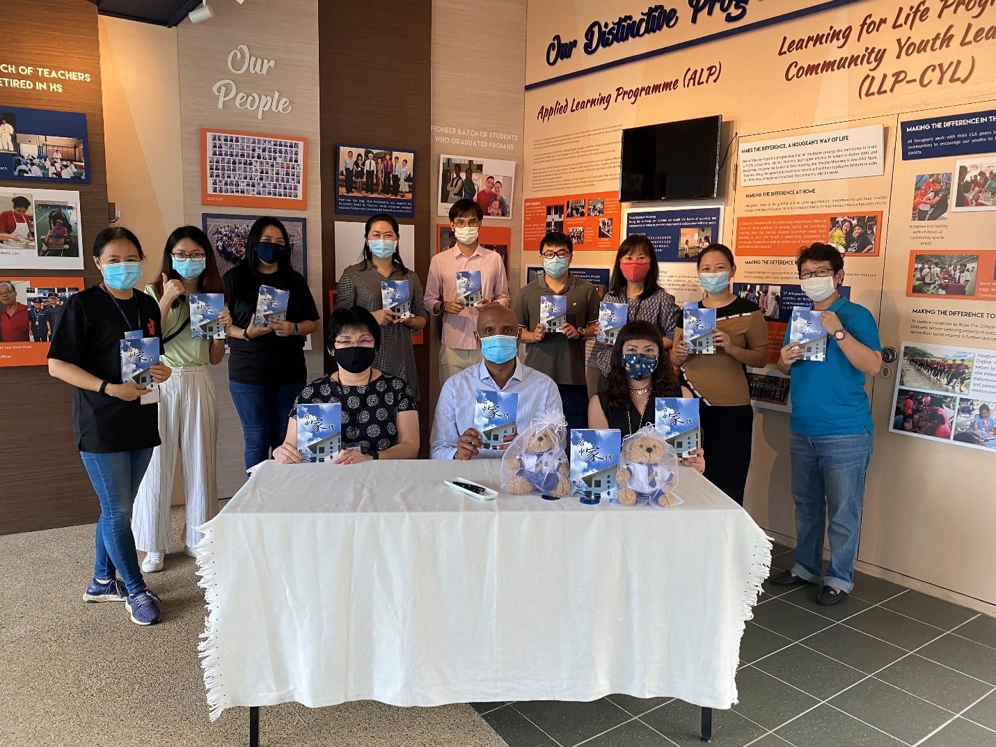 A group of masked people holding blue booklets pose in a school exhibition hall with informational displays.