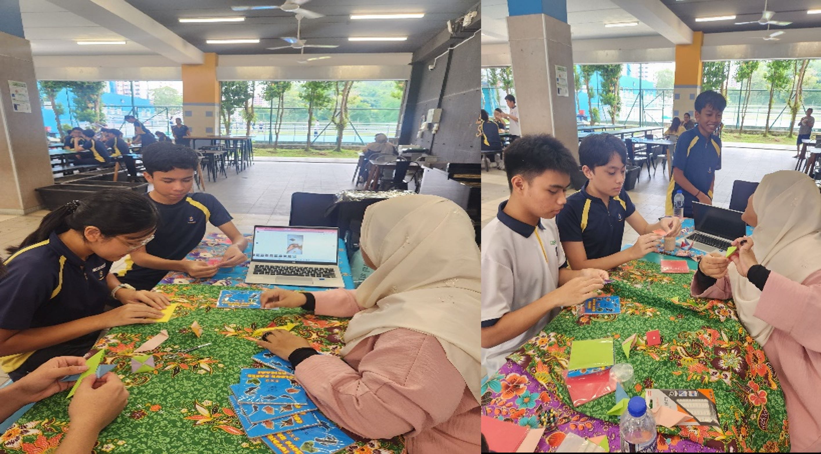 Students in uniforms and a woman in a hijab fold origami paper at a table with a laptop in an open hall.