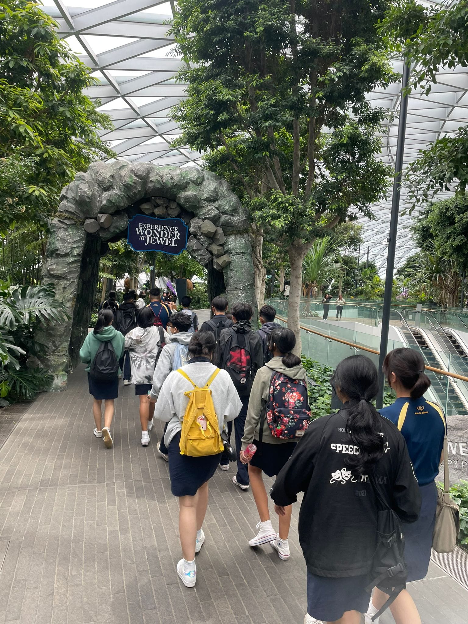 Students with backpacks walk toward a stone arch sign reading "Experience Wonder at Jewel" inside a lush indoor garden.