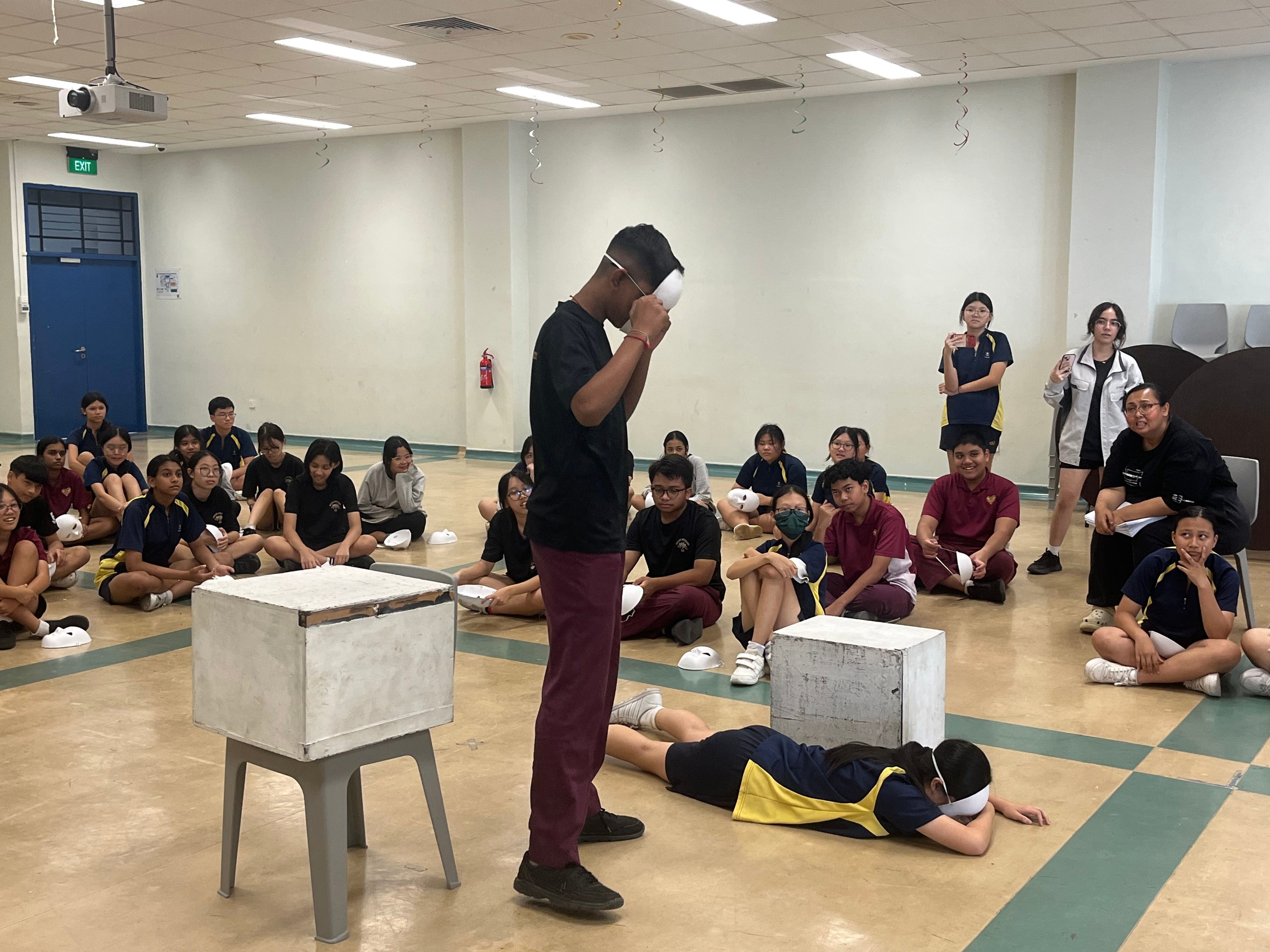 A student wearing a white mask stands in a hall while others sit on the floor watching during a school activity.