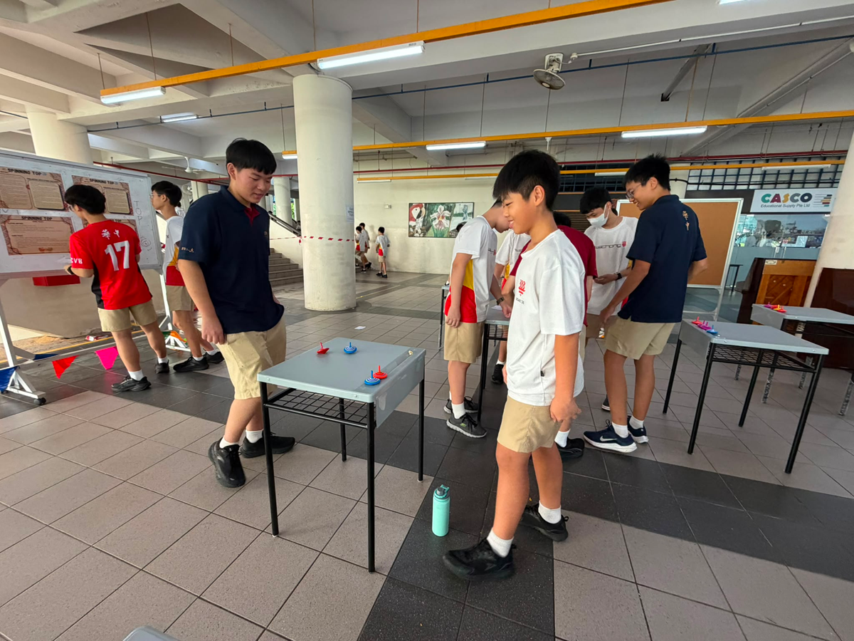 High school students gathered around small desks in the school's sheltered plaza trying out traditional spinning tops (Gasing) at a heritage game station.