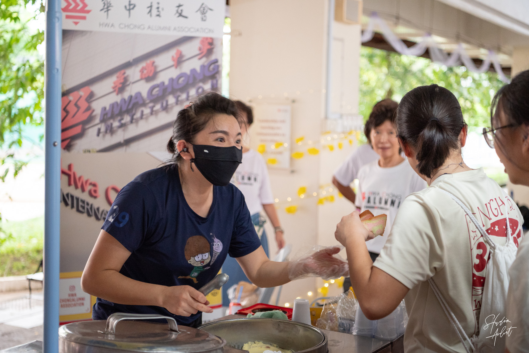 A masked woman in a navy shirt serves food at a Hwa Chong Institution alumni event booth to waiting customers.
