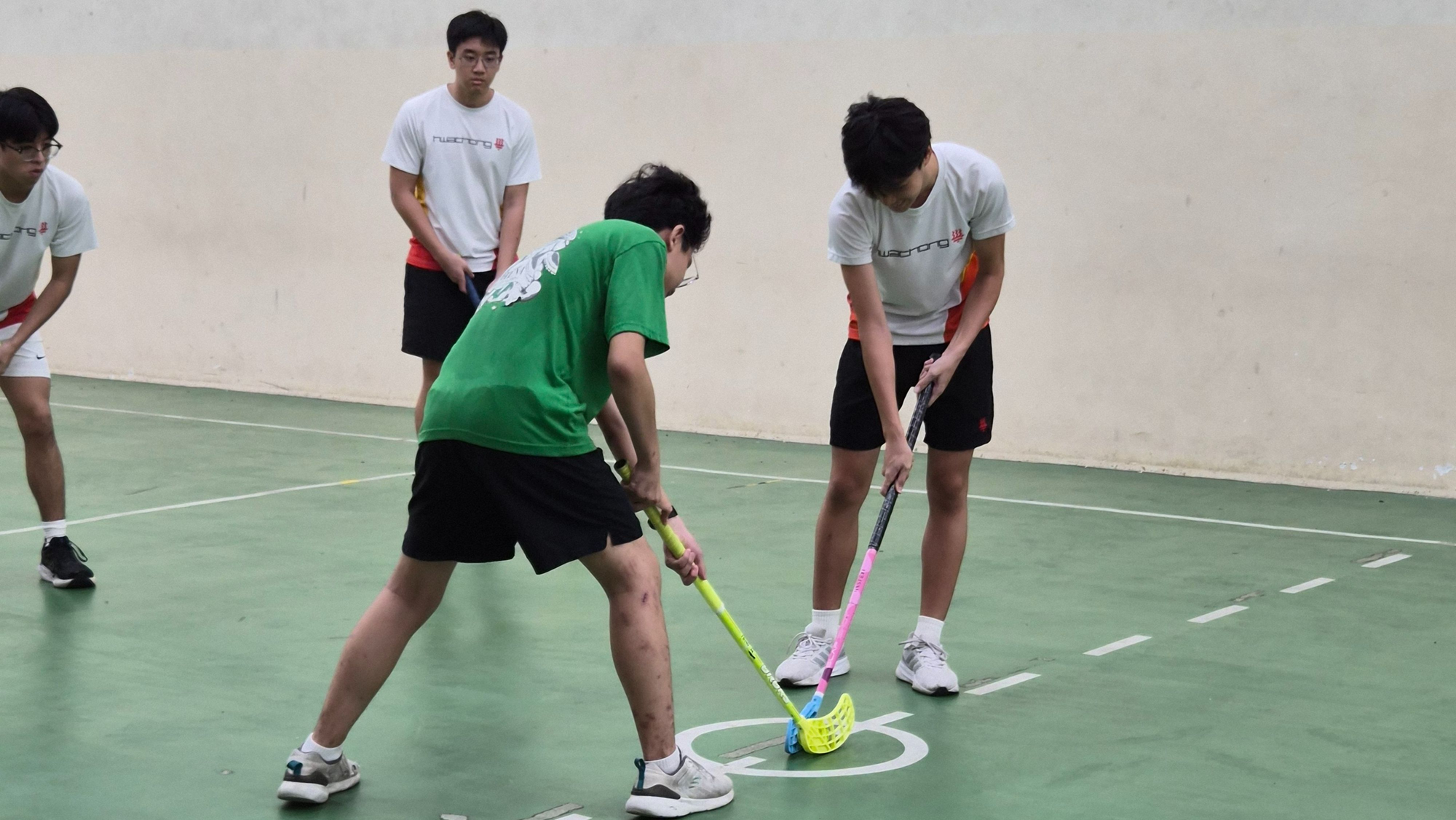 Four young men play floorball indoors, two competing for the ball with colorful sticks on a green court.