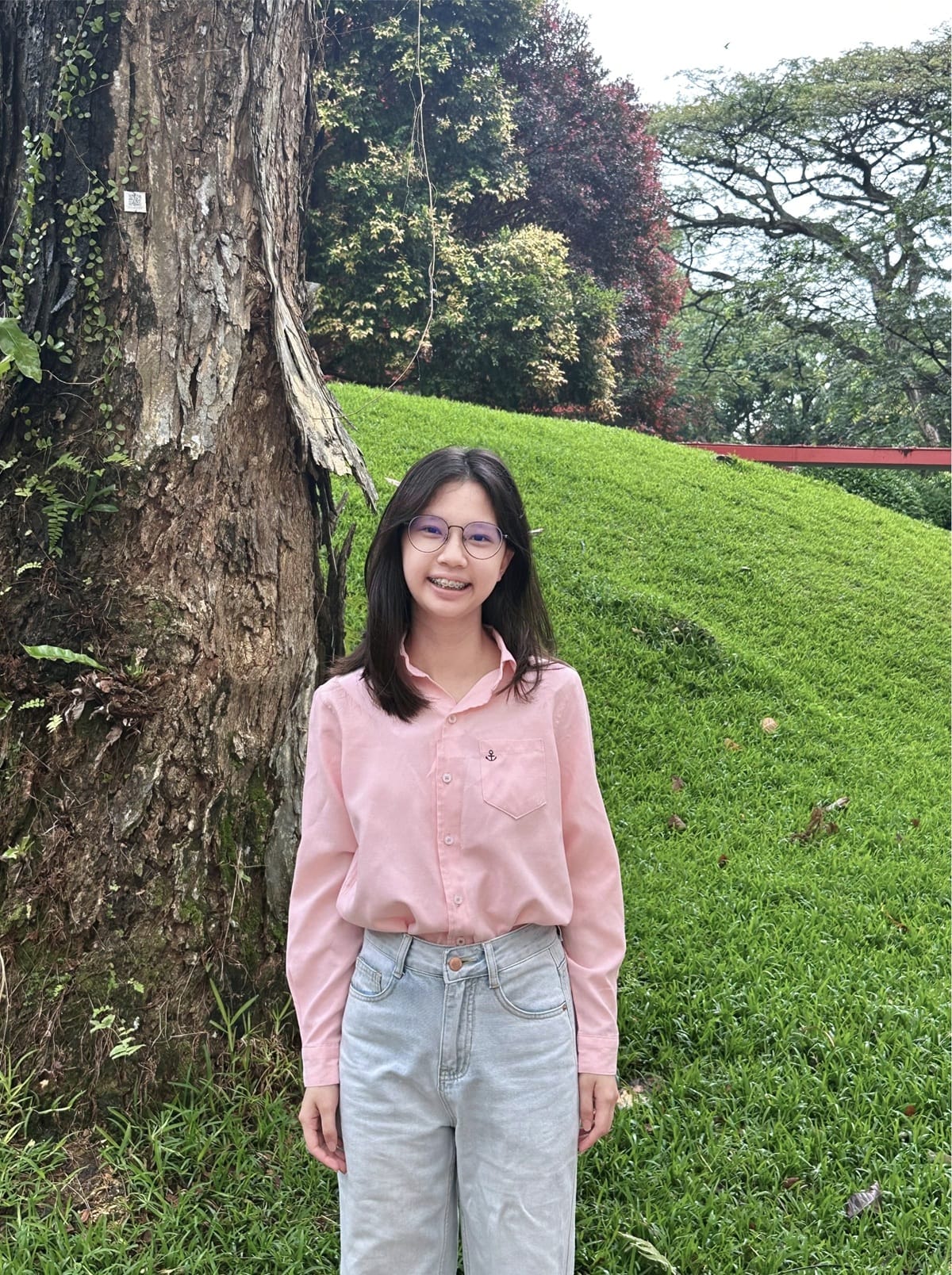 Young woman with glasses and braces in a pink shirt and jeans smiling beside a large tree on a green grassy hill.