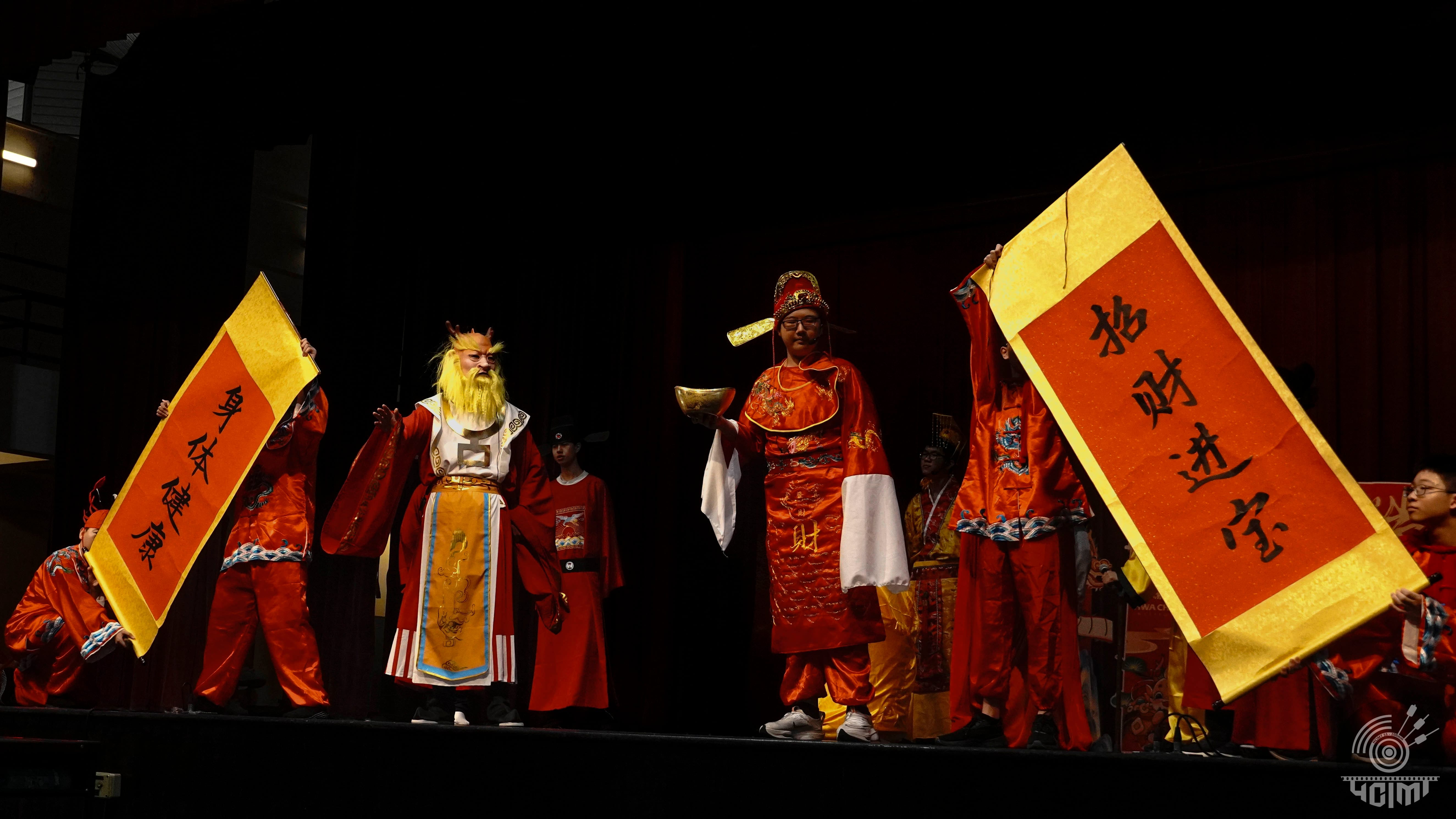 Performers in red traditional Chinese costumes hold large yellow banners with Chinese characters on a dark stage.