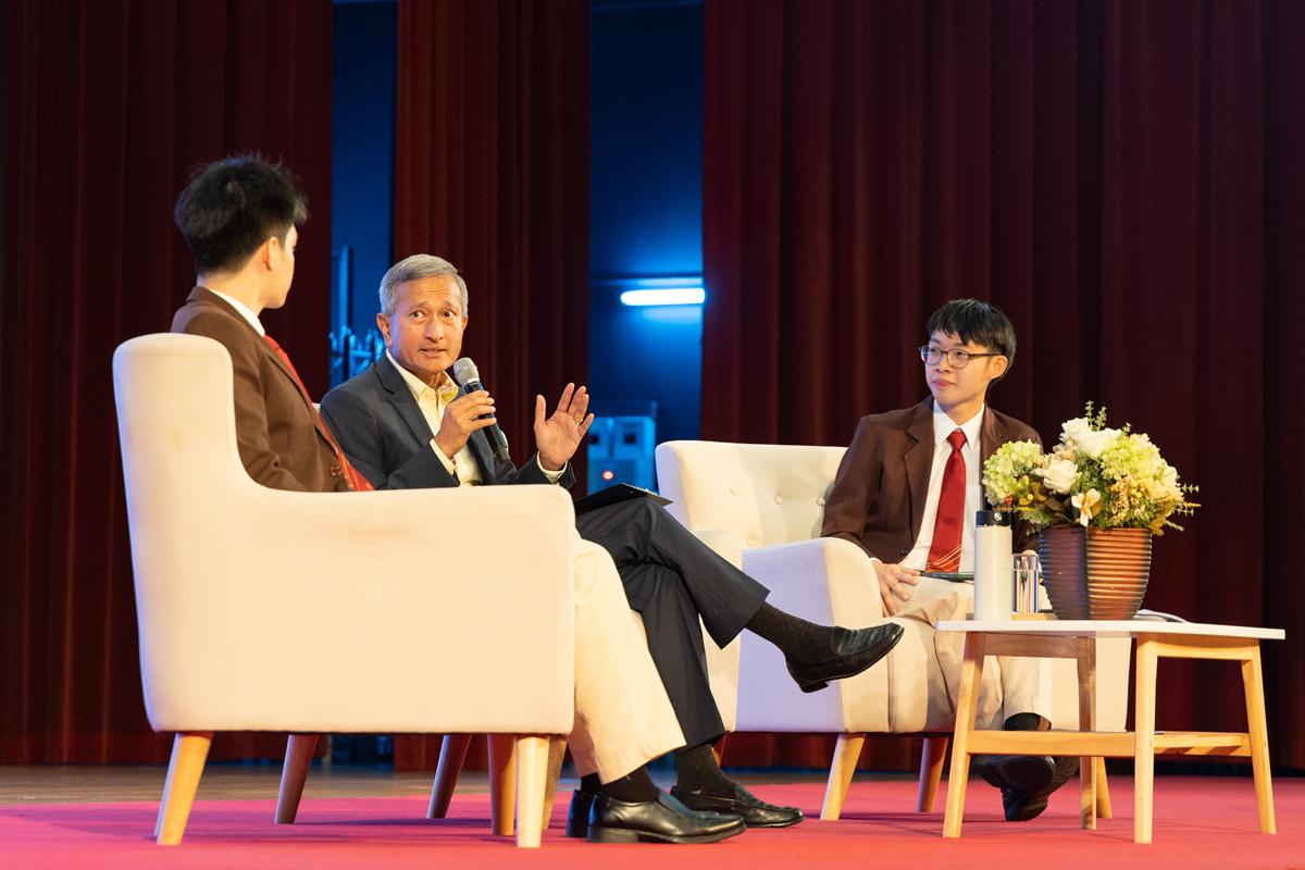 Three people sit in white armchairs on a stage, with a man in a suit speaking into a microphone before a red curtain.