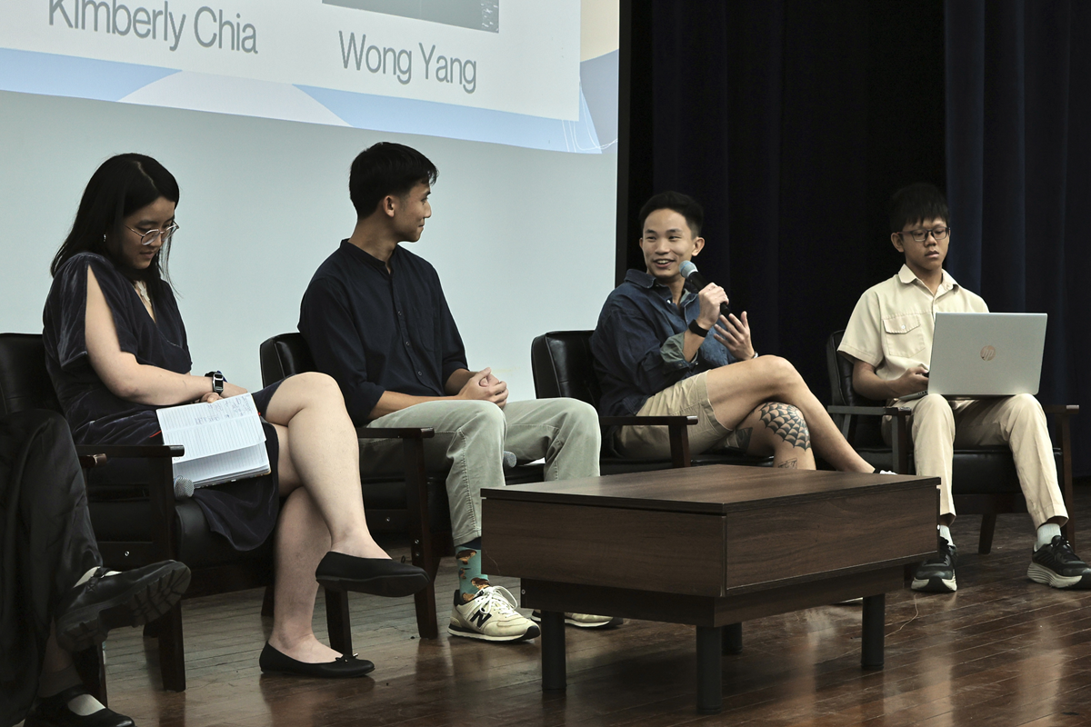 Four panelists seated on stage, one speaking into a microphone, with a projected slide showing names Kimberly Chia and Wong Yang.
