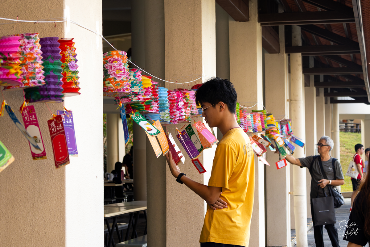 A male student in a yellow shirt reading a lantern riddle slip hanging along a sheltered walkway decorated with colorful paper lanterns during the Mid-Autumn Festival.