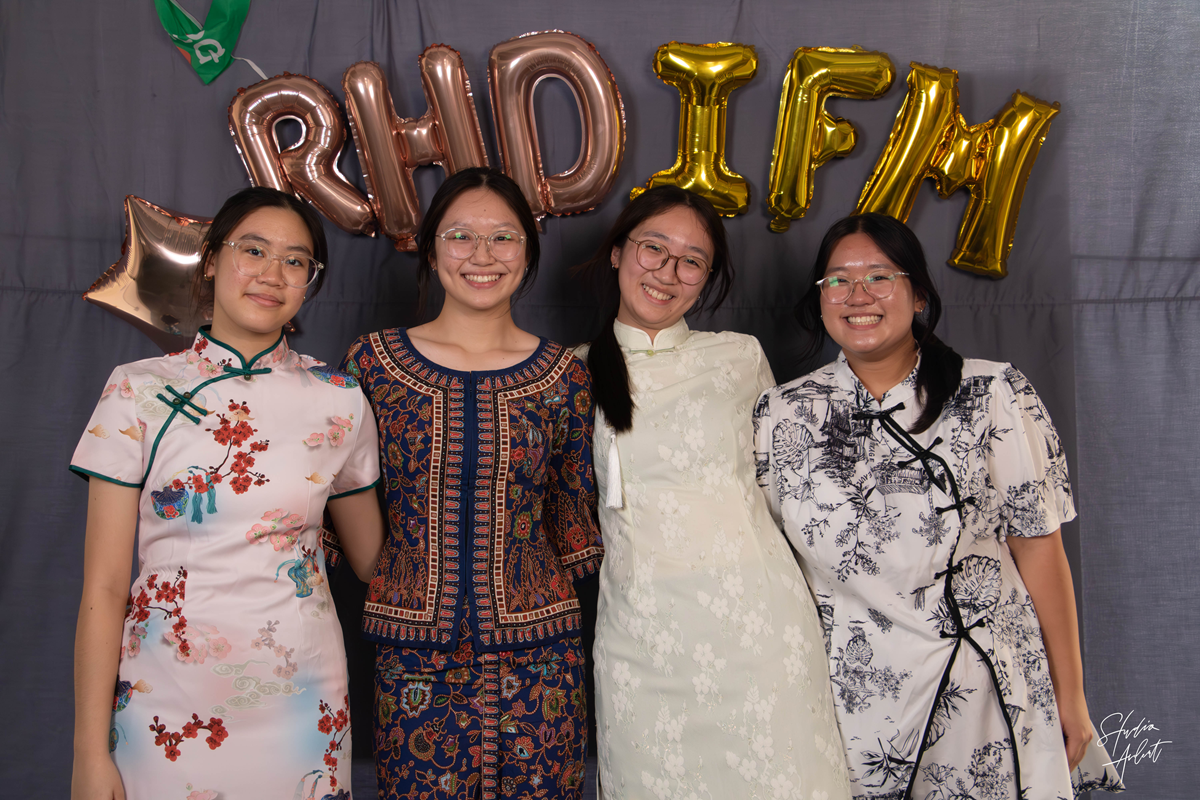 Four smiling Hwa Chong Institution students standing side-by-side in front of a grey backdrop with metallic foil balloons spelling "RHD IFM". They are dressed in diverse traditional attire, including Cheongsams and a Kebaya.