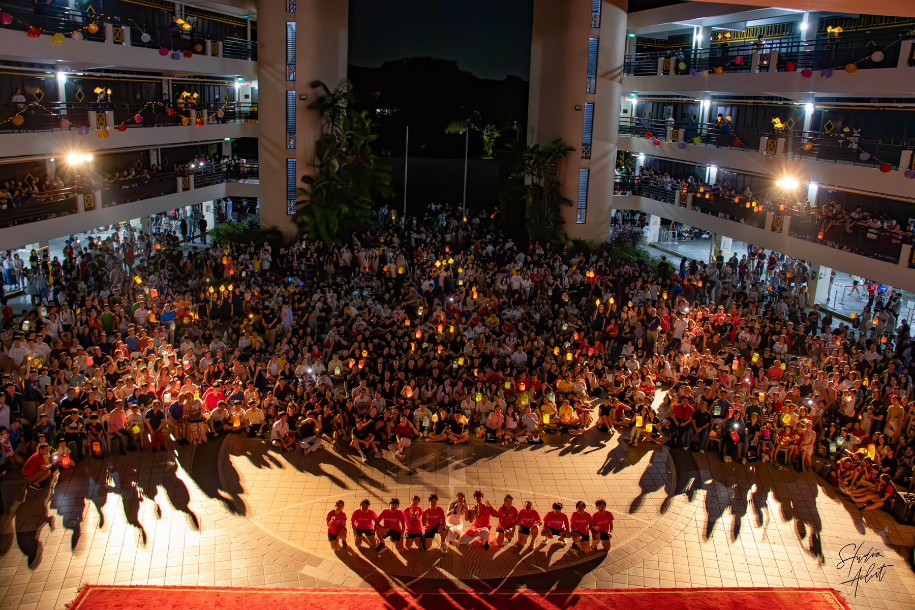 Large crowd fills a multi-level atrium, facing a seated group in red shirts under bright stage lights.