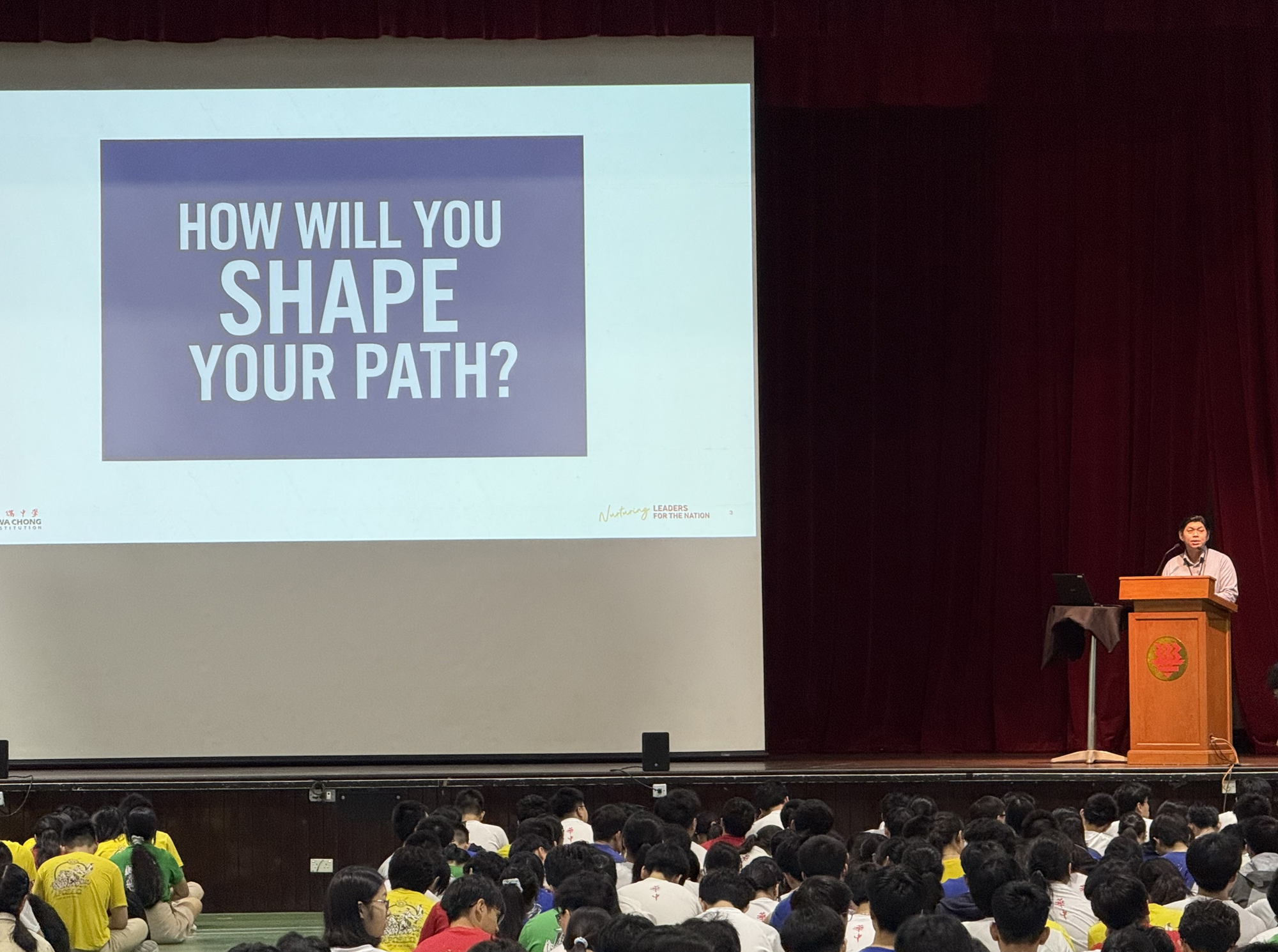 A speaker at a podium presents a slide reading "How Will You Shape Your Path?" to a large student audience in an auditorium.