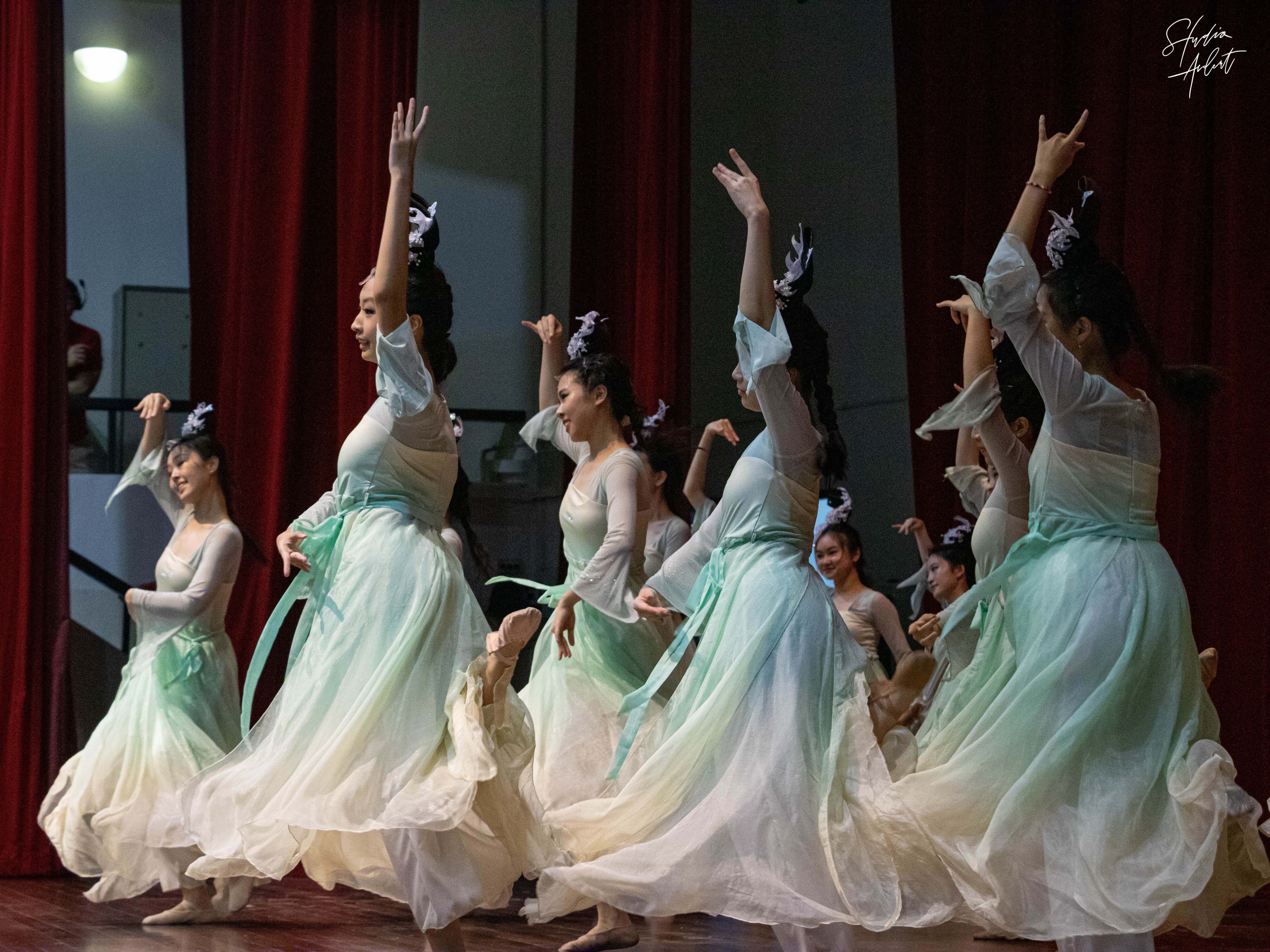 Several women in flowing green and white traditional Chinese dresses perform a synchronized dance on a stage.