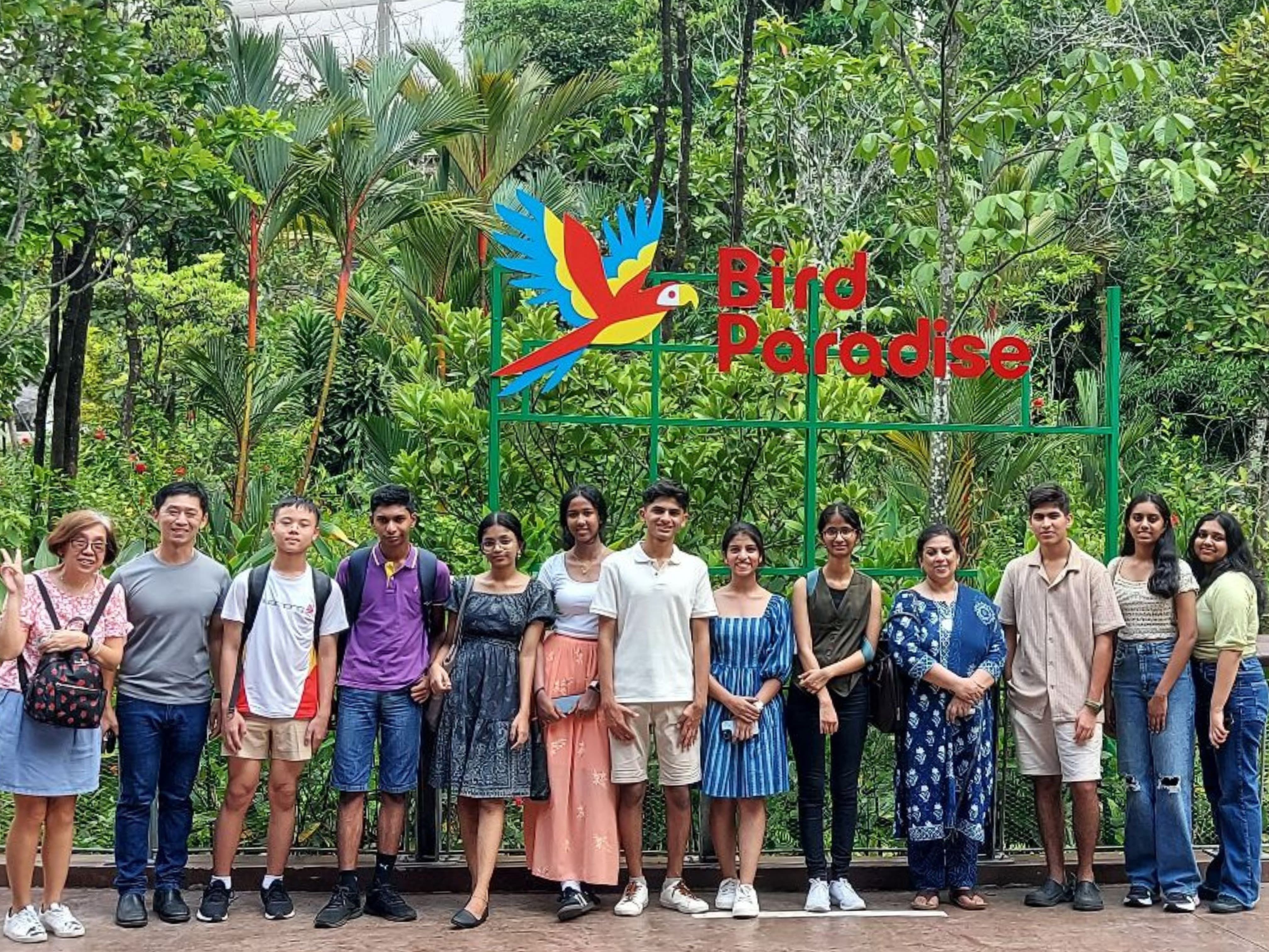 A group of about 13 people posing in front of the Bird Paradise sign with a colorful parrot mascot and green trees.