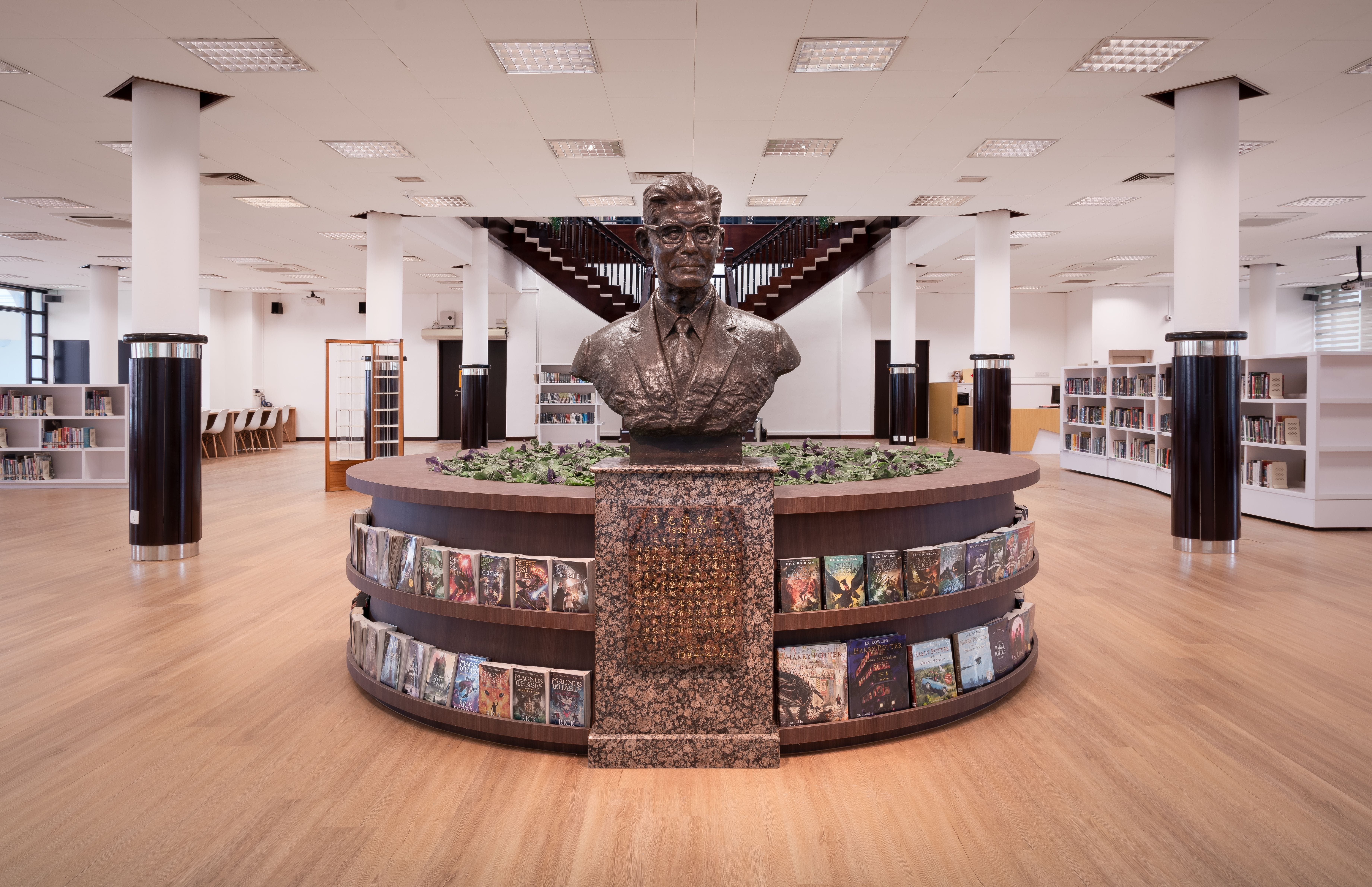 Bust statue on a circular book display in a bright library, with stairs and shelves in the background