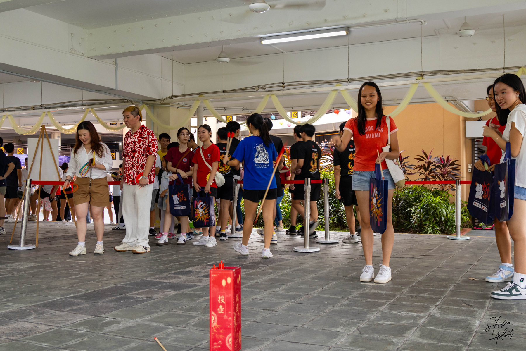 A crowd of people queue behind red rope barriers at a festive Asian event with decorations and a red lantern on the floor.