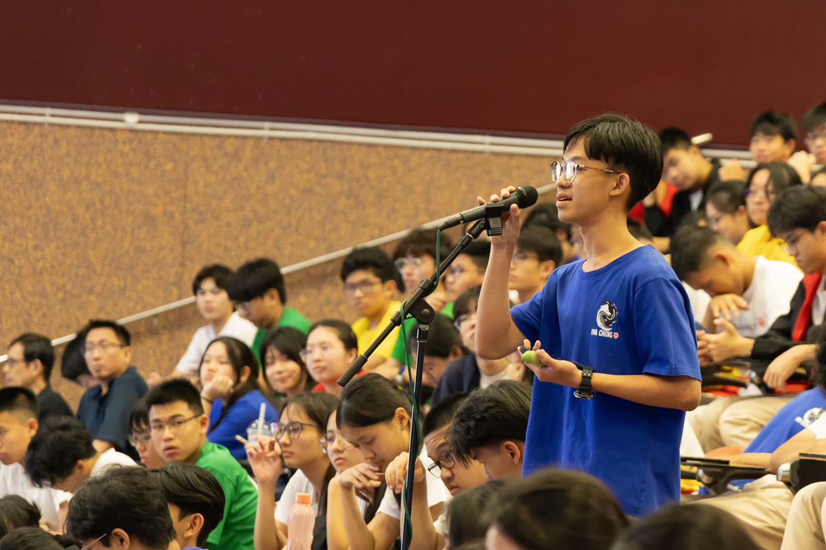 A young male student in a blue shirt speaks into a microphone before a large seated audience.