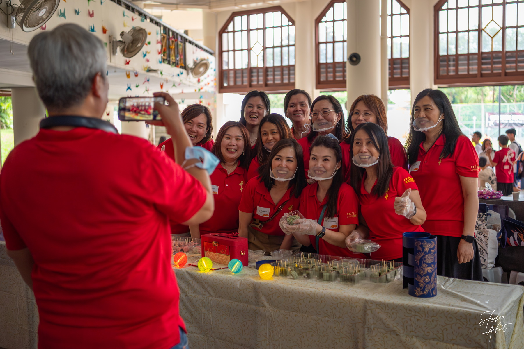 A person photographs a group of smiling women in red polo shirts posing behind a table with small plants and colorful lanterns.