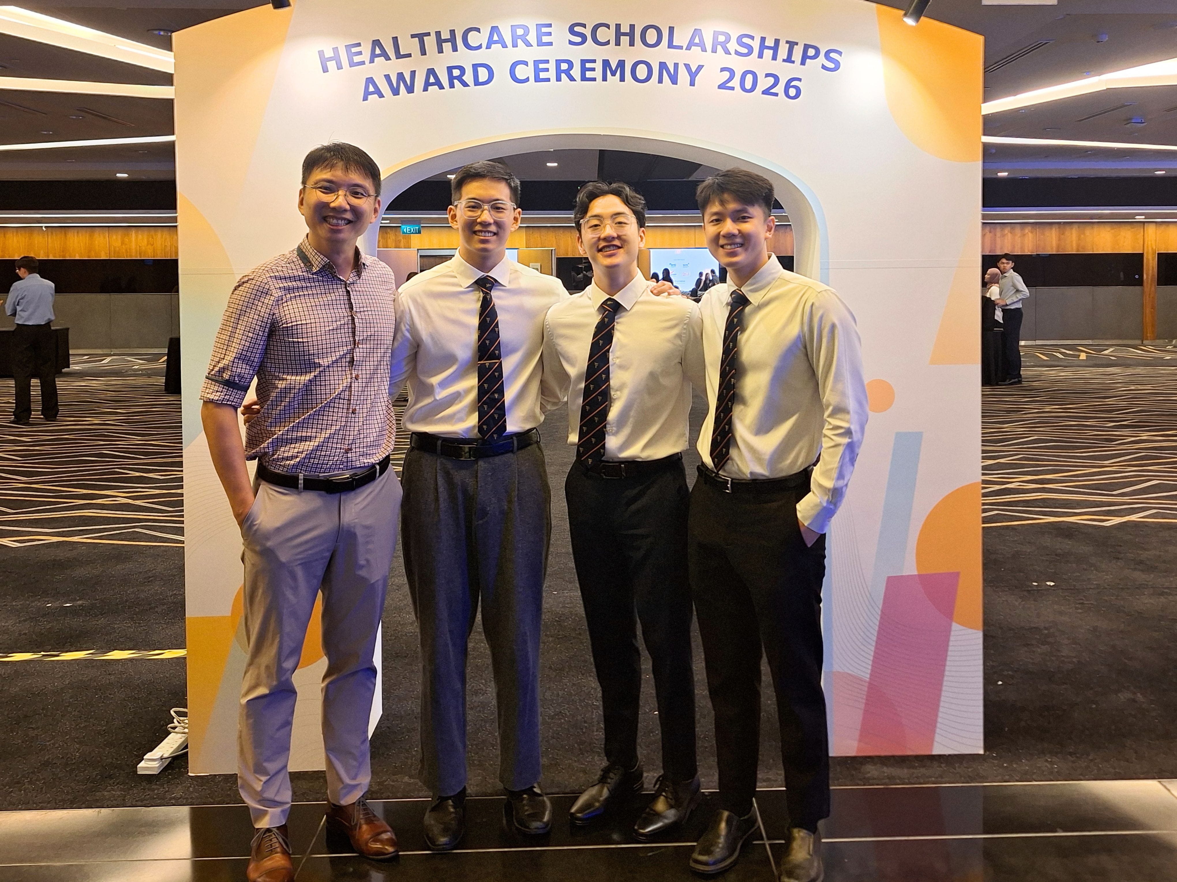 Three male scholars in formal shirts and school ties pose with a male teacher in front of the Healthcare Scholarships Award Ceremony 2026 backdrop.