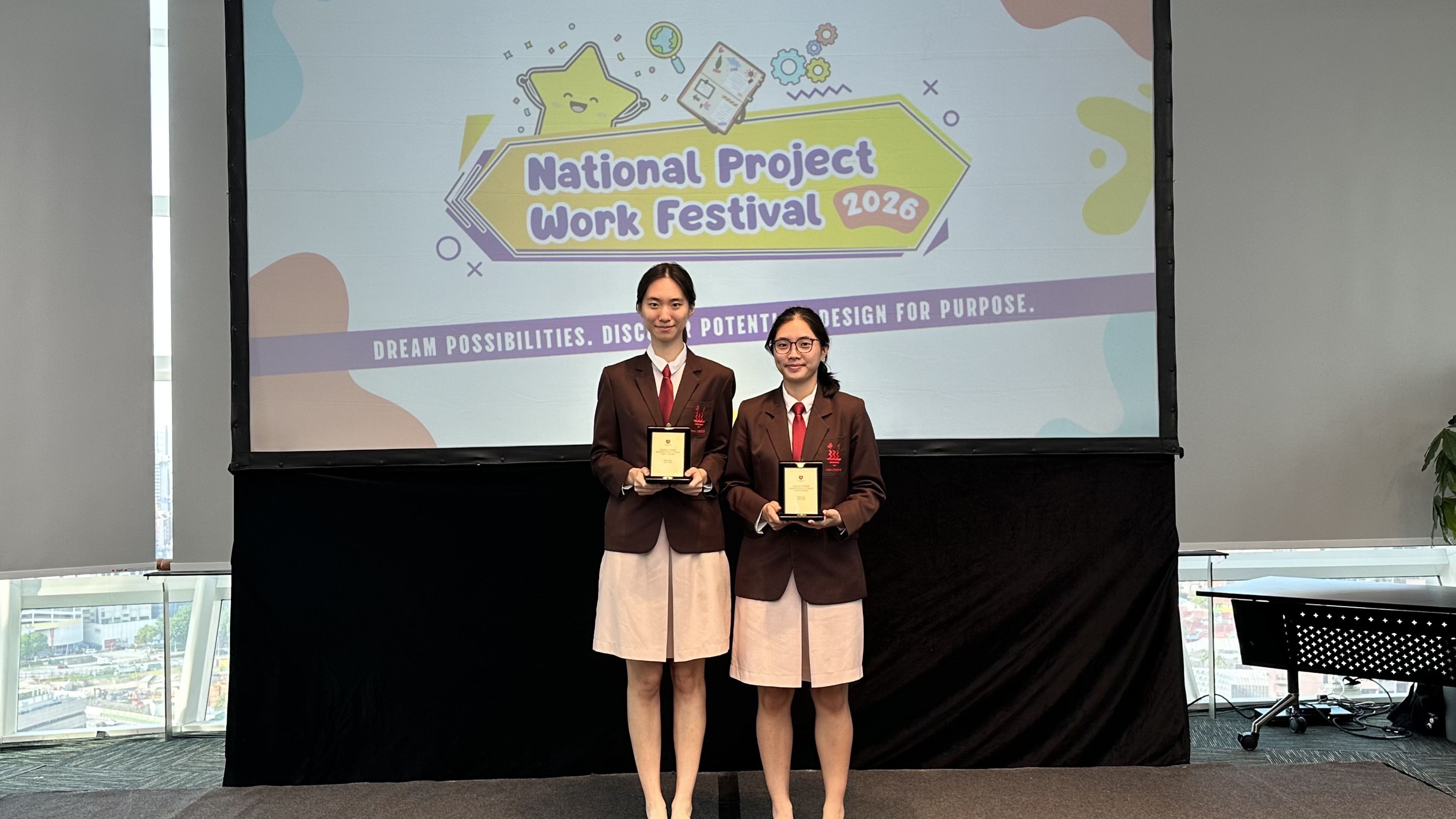 Two female students in brown blazers hold awards in front of a National Project Work Festival 2026 banner.