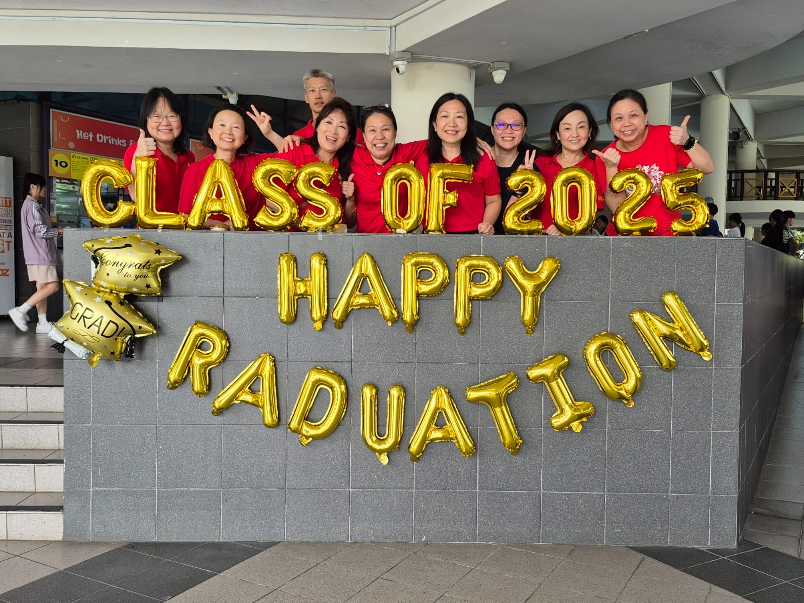 Group of people in red shirts posing behind gold balloon letters reading Class of 2025 Happy Graduation.