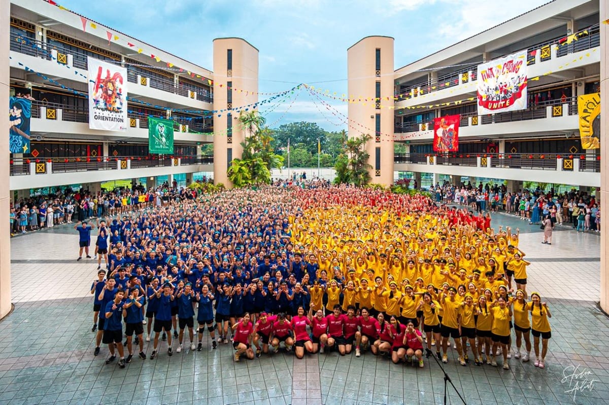 Large school group photo in a courtyard, students in blue, yellow, red, and pink shirts posing cheerfully.