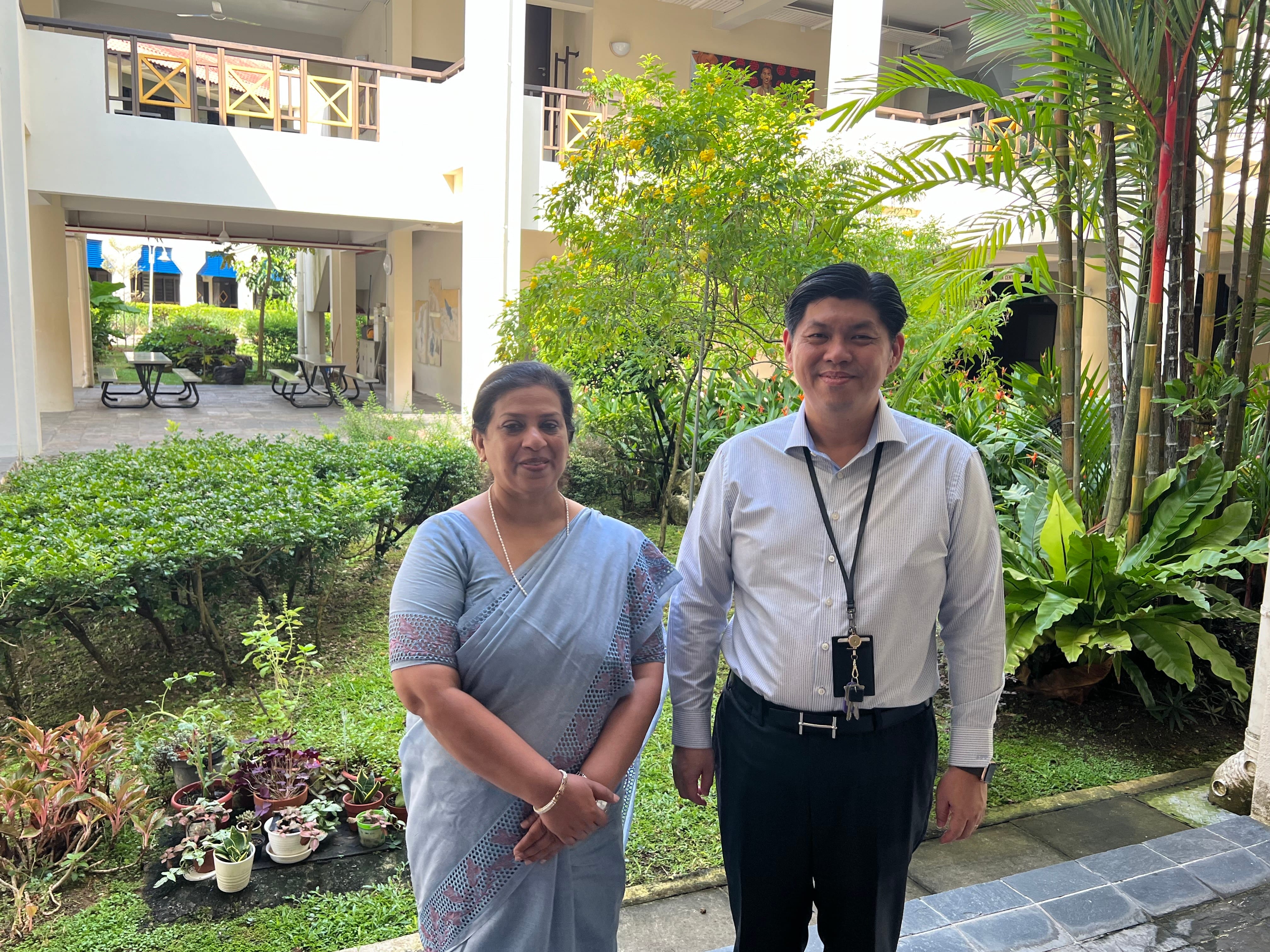 Two people stand smiling outdoors in a lush garden courtyard; a woman in a blue sari and a man in a white shirt with a lanyard.