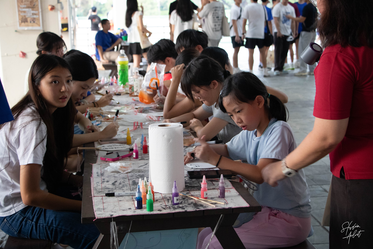 Students seated along an outdoor wooden bench participating in a craft workshop with paints and markers during the festival.