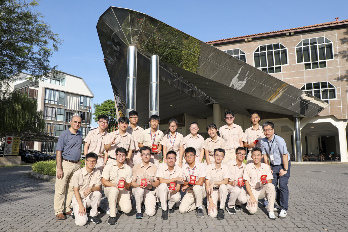 A group of students in beige uniforms holding red medal boxes pose with two adults outside a modern building.