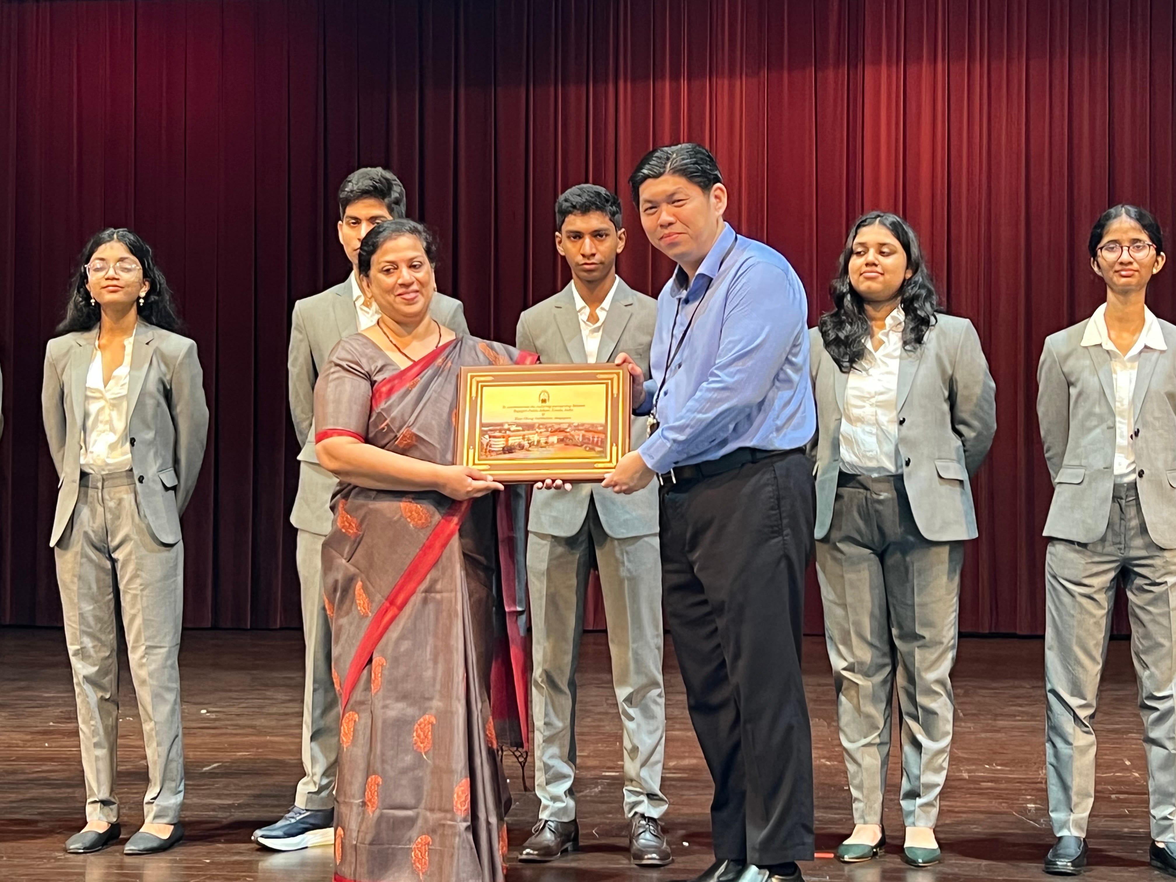 A woman in a saree and a man in a blue shirt jointly hold a framed plaque on stage, with students in grey suits standing behind.