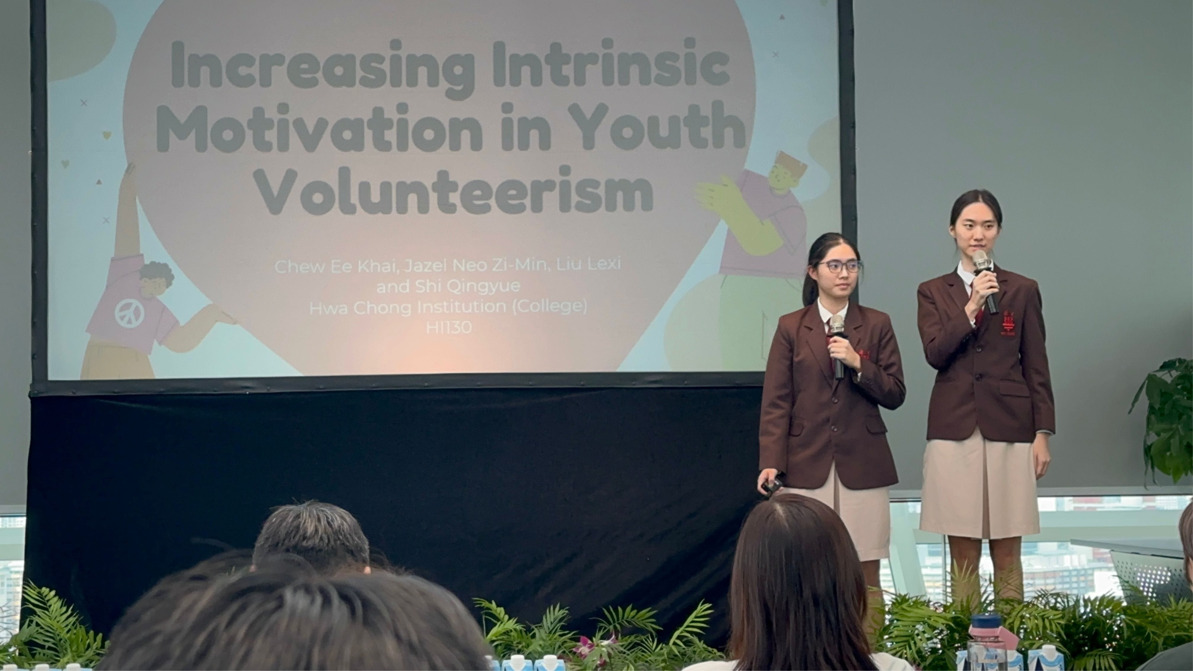 Two students in brown uniforms hold microphones presenting on "Increasing Intrinsic Motivation in Youth Volunteerism."