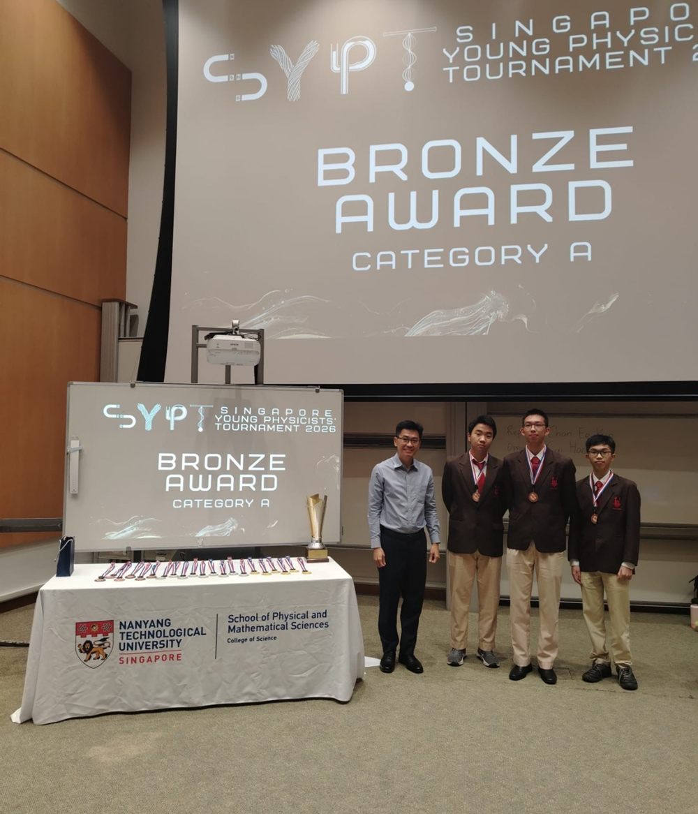 Four people stand beside a table with medals at the Singapore Young Physicists Tournament Bronze Award Category A ceremony.
