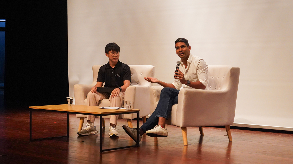 Mr. Gaurav Keerthi speaking into a microphone while seated in an armchair on stage next to a student facilitator.