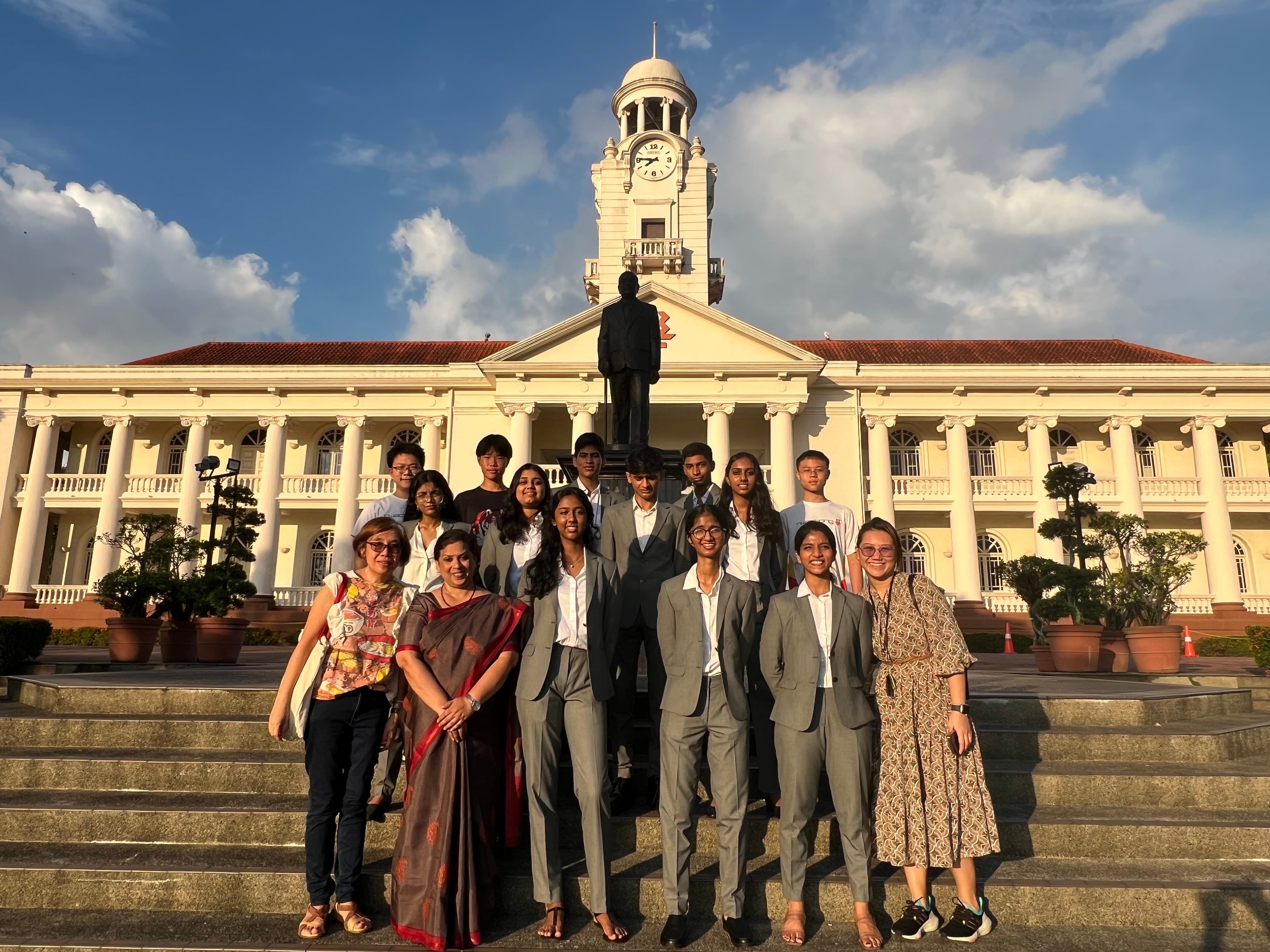 Group of students and adults posing on steps before a white colonial building with a clock tower and statue.