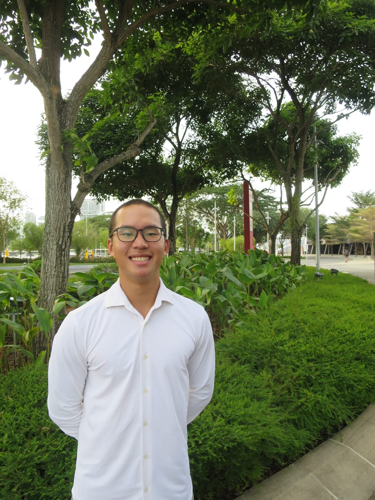 Young man with glasses and a white shirt smiling in a green park with trees and trimmed hedges behind him.