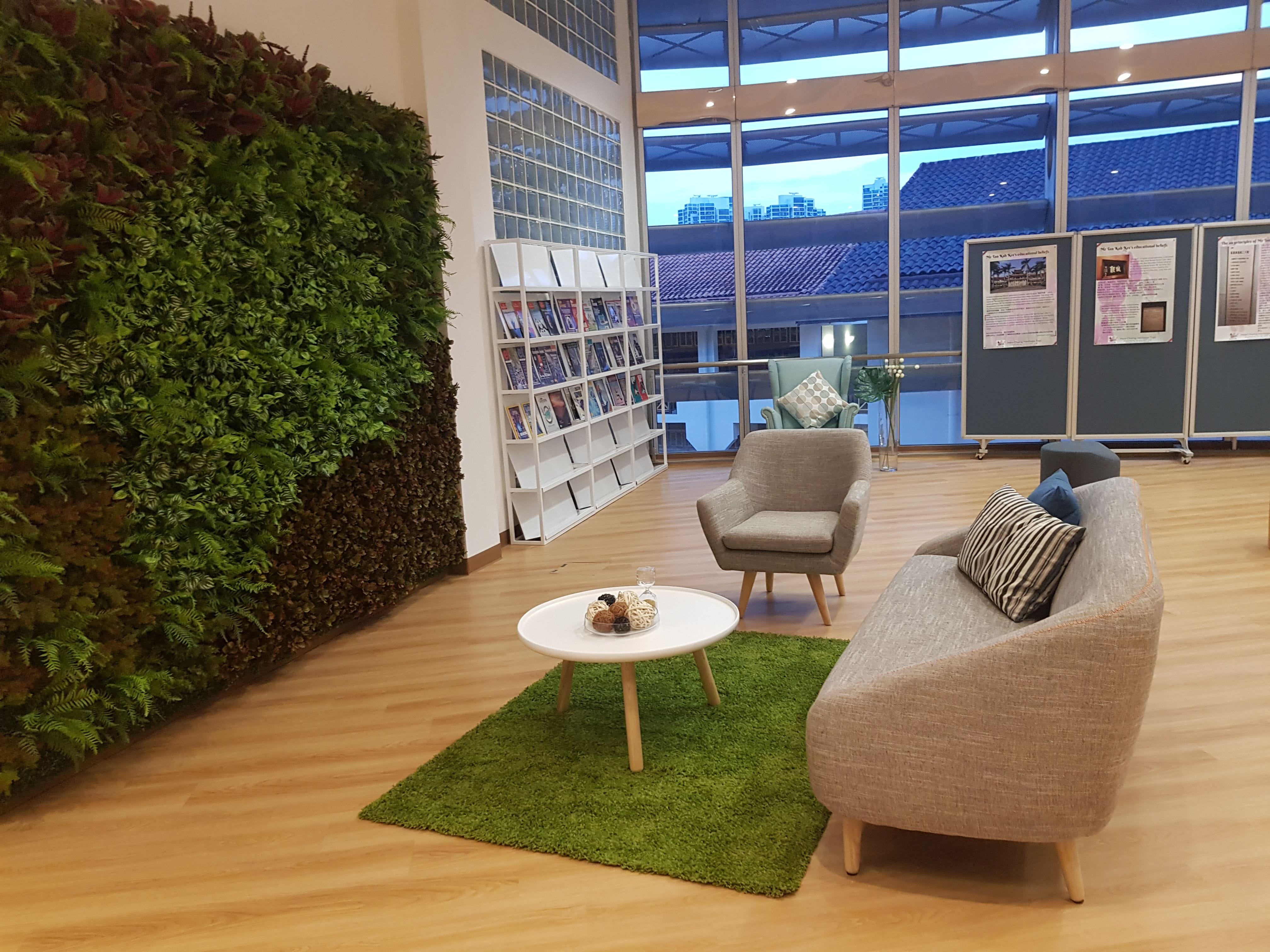 Modern lounge with a green living wall, gray sofa and chair, white round coffee table on a green rug, and a magazine rack.